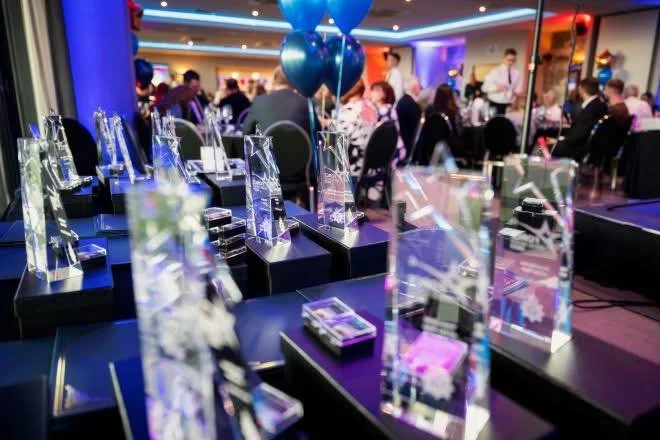 Display of clear acrylic awards on black stands at a crowded indoor event with balloons and people in the background.