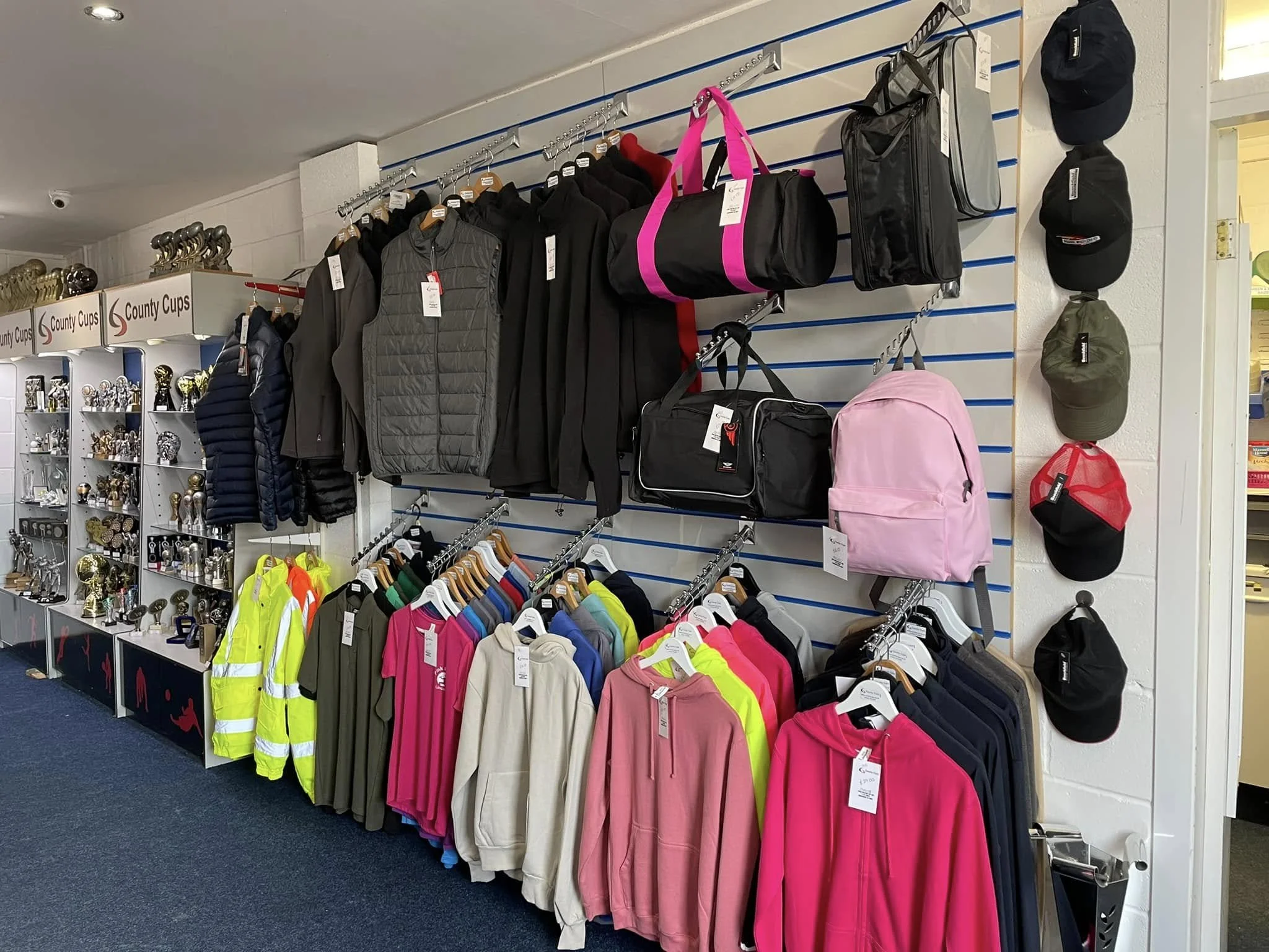 Clothing and accessories display in a retail store, with jackets, hoodies, and hats on a wall with hooks, and high-visibility safety vests and various trophies on shelves to the side.