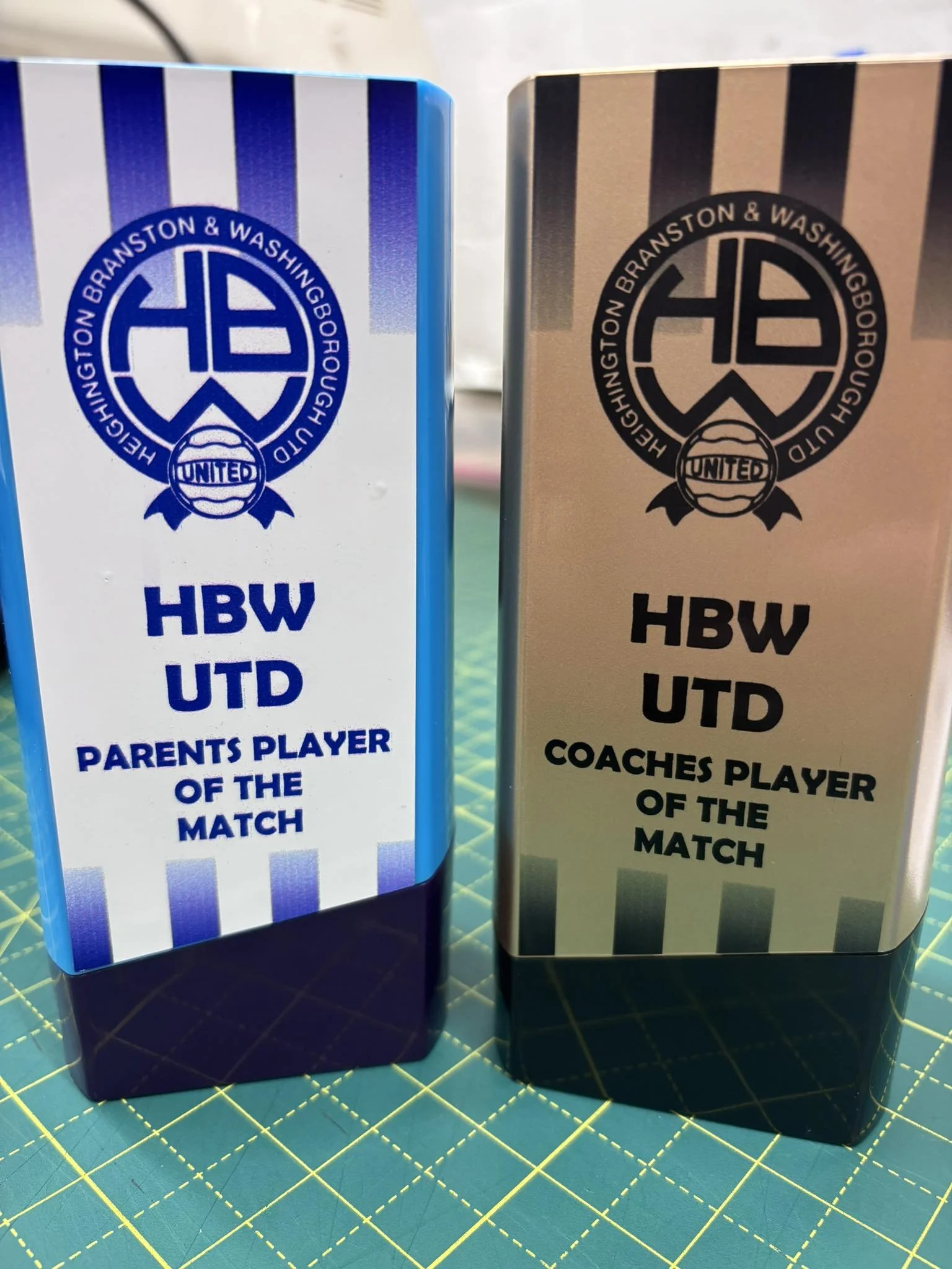 Two rectangular awards with the HEBU logo, one with blue and white stripes and the other with black and silver stripes, labeled for best parents and coach players of the match.