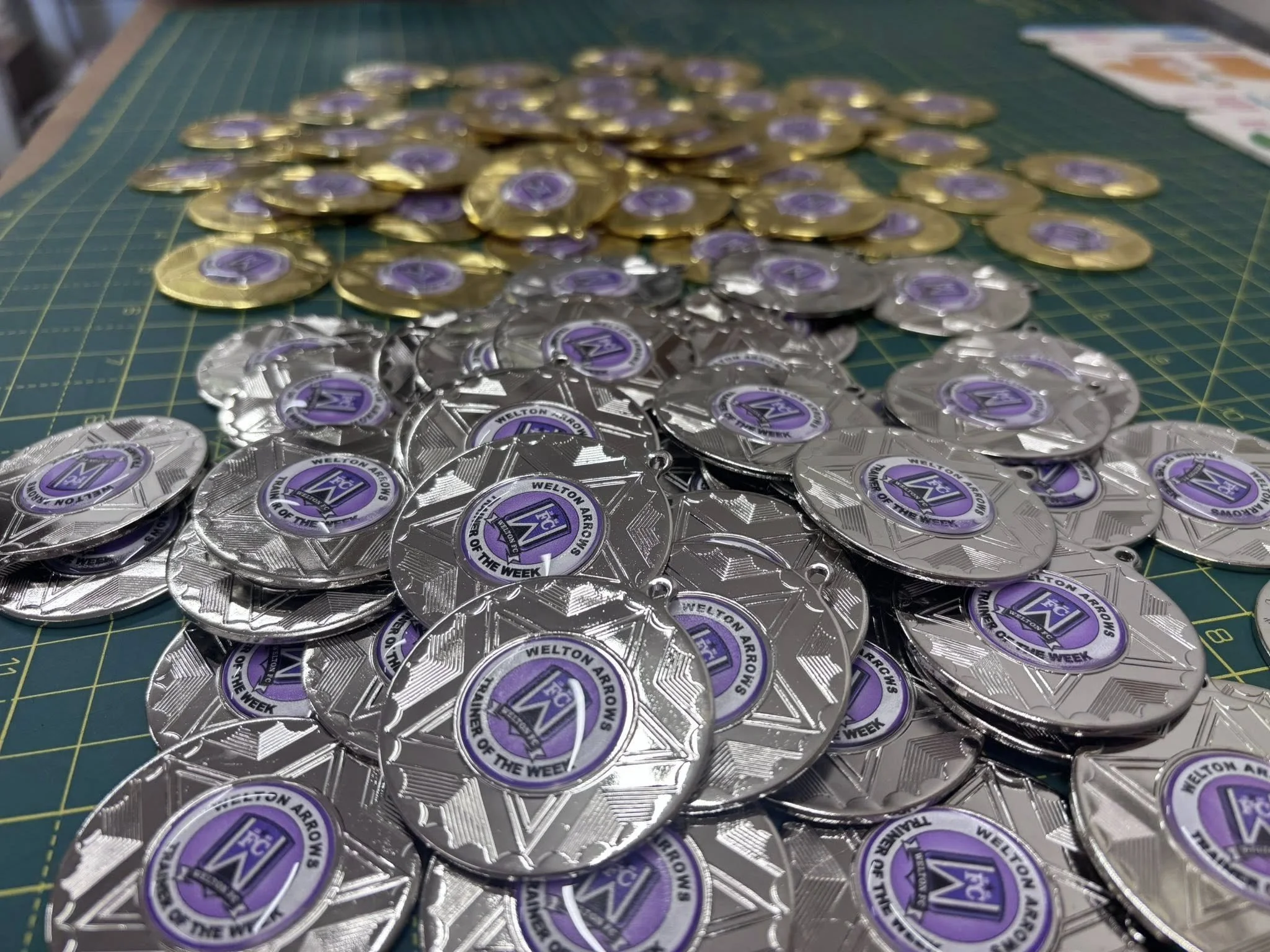 Silver and gold medals with purple and black emblem, stacked on a green cutting mat.
