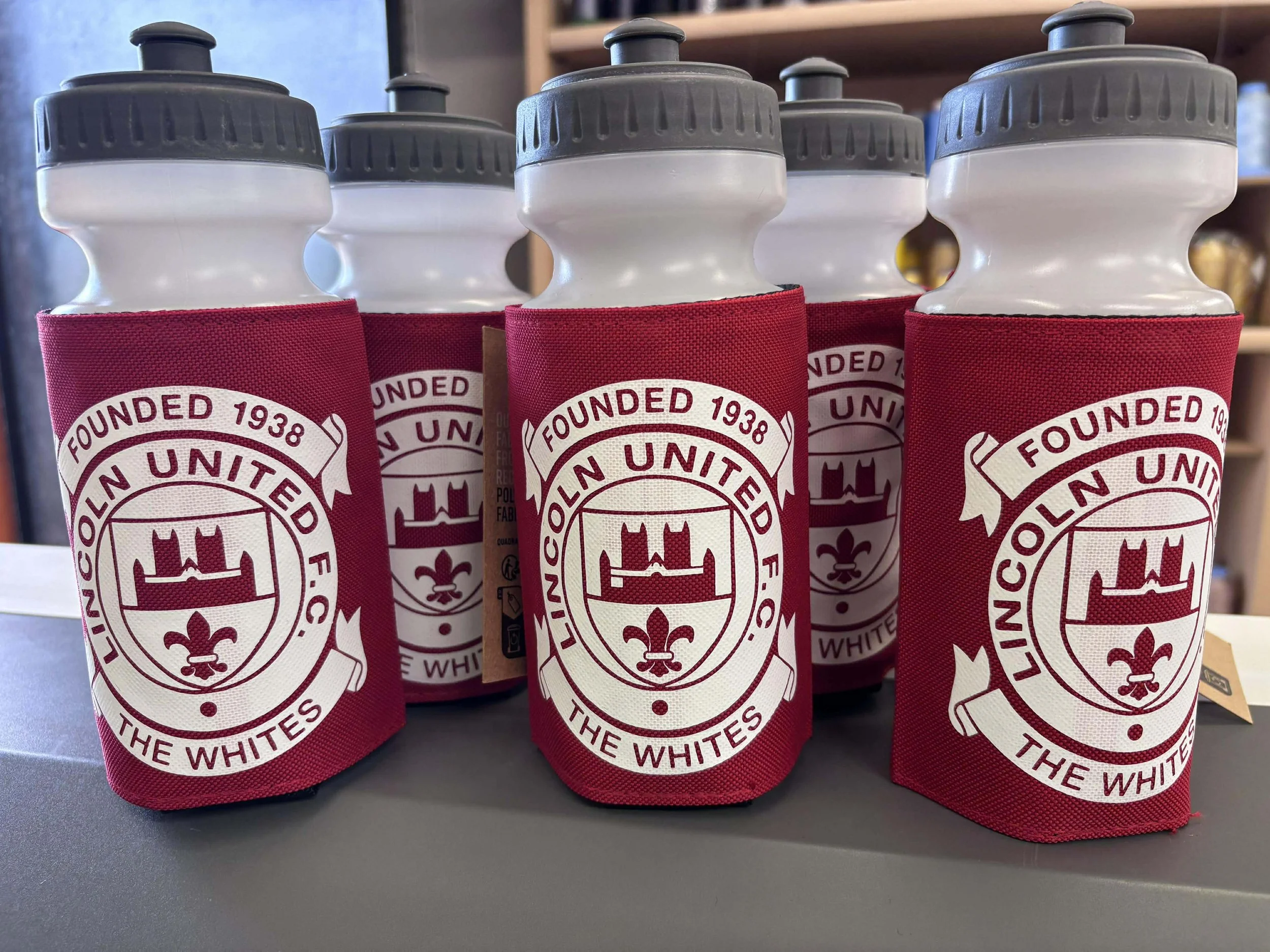 Five water bottles in red fabric holders with the Lincoln United F.C. logo and text, on a gray surface with a background of shelves.