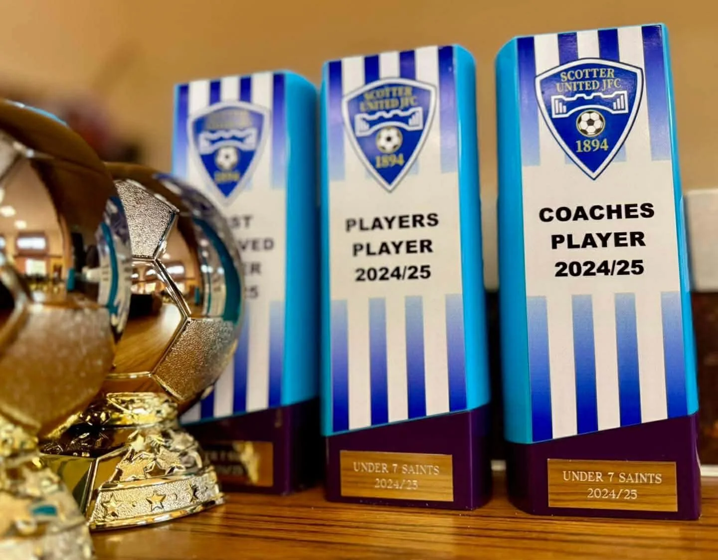 Miniature gold soccer ball trophy, with a reflective surface, next to two blue and white team program guides labeled 'Players Player 2024/25' and 'Coaches Player 2024/25' for the Under 7 Saints soccer team.