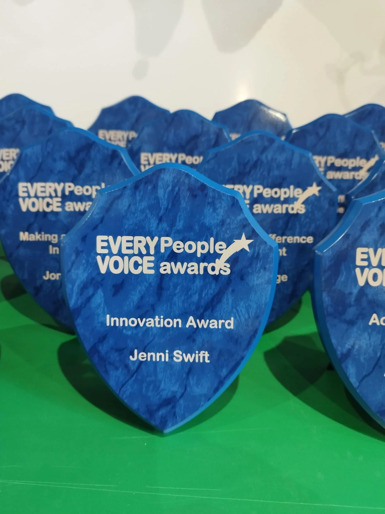 Blue award trophy shaped like a shield with white text reading 'Every People Voice Awards', 'Innovation Award', and 'Jenni Swift.'