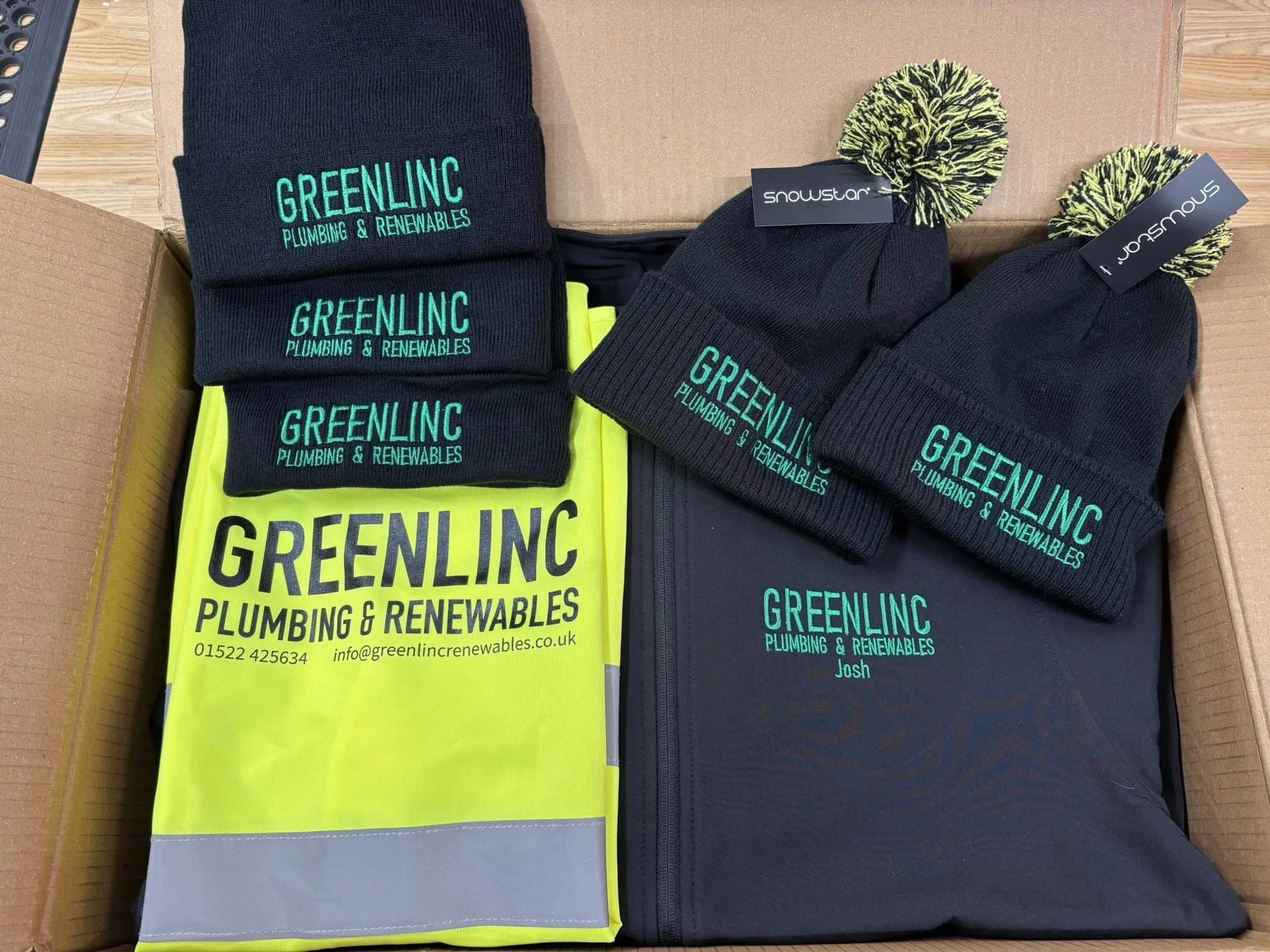 A box containing clothing items with custom branding for Greenlinlc Plumbing & Renewables. Inside are three beanie hats, two with yellow and black pom-poms and one with a plain black design, all with the Greenlinlc logo. Also included is a high-visibility safety vest with the Greenlinlc logo and contact information, and a black jacket with the Greenlinlc logo and the name "Josh."