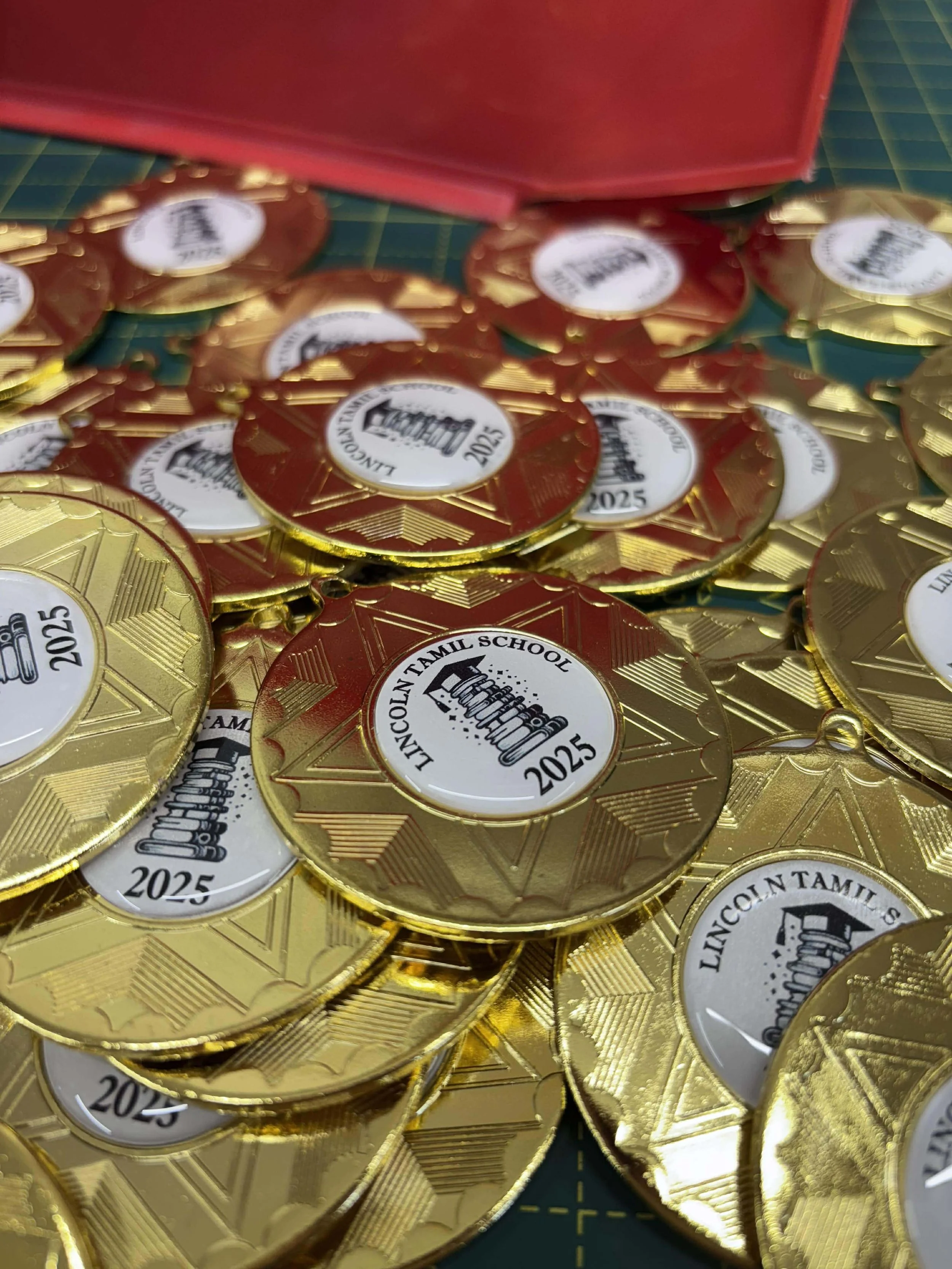 A pile of gold and red medals with a white center that reads 'Lincoln Tamil School 2025' and features a drawing of bowling pins.