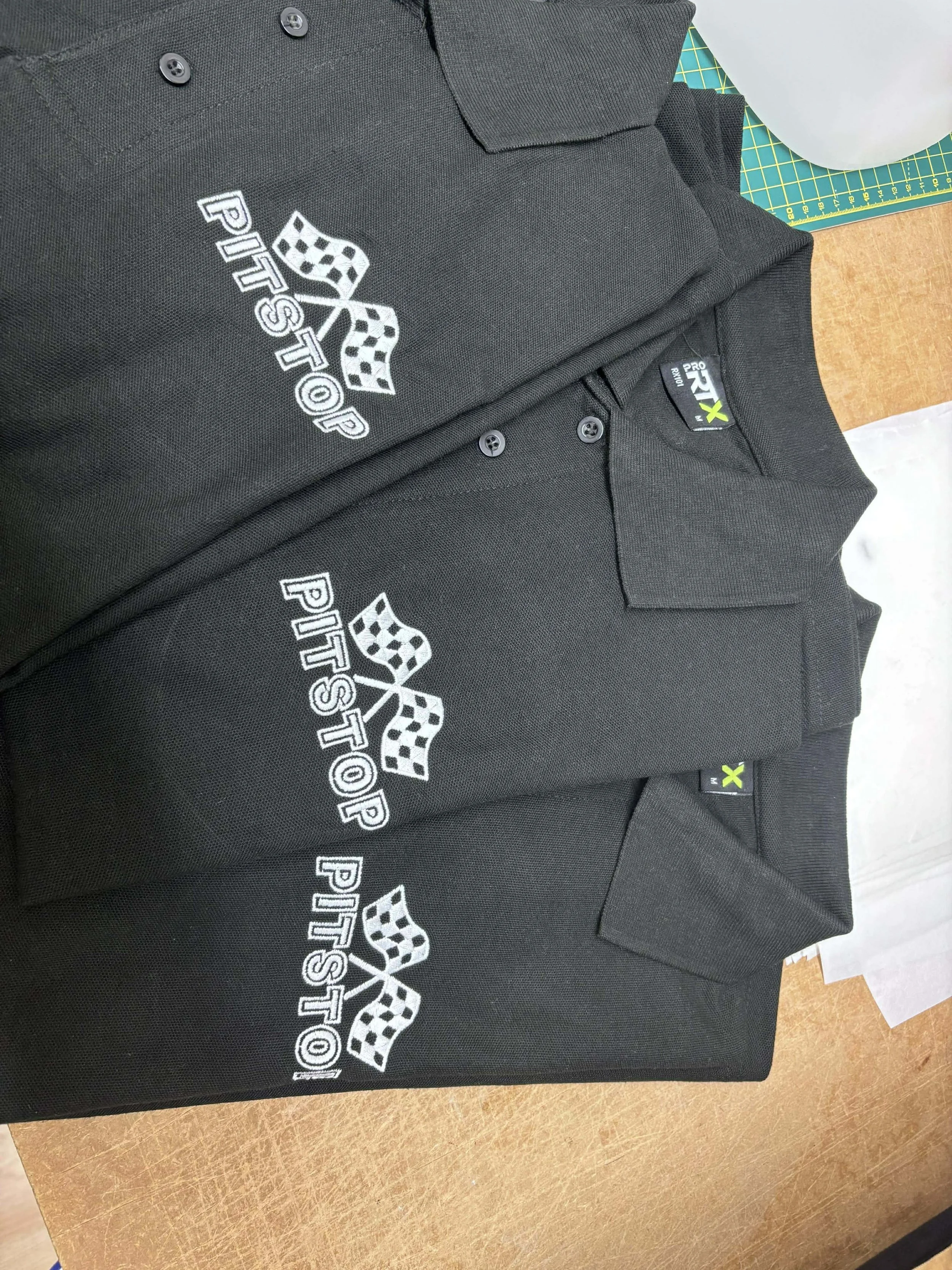 Three black polo shirts with white and checkered flag embroidered patches on the chest, laid out on a wooden surface with a green cutting mat in the background.