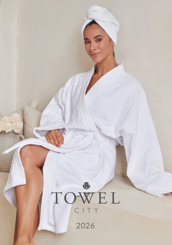 A woman in a white bathrobe and towel wrapped around her head, sitting on a cushioned bench against a neutral wall, with a small bouquet of flowers in a beige pillow nearby, promoting Towel City 2026.