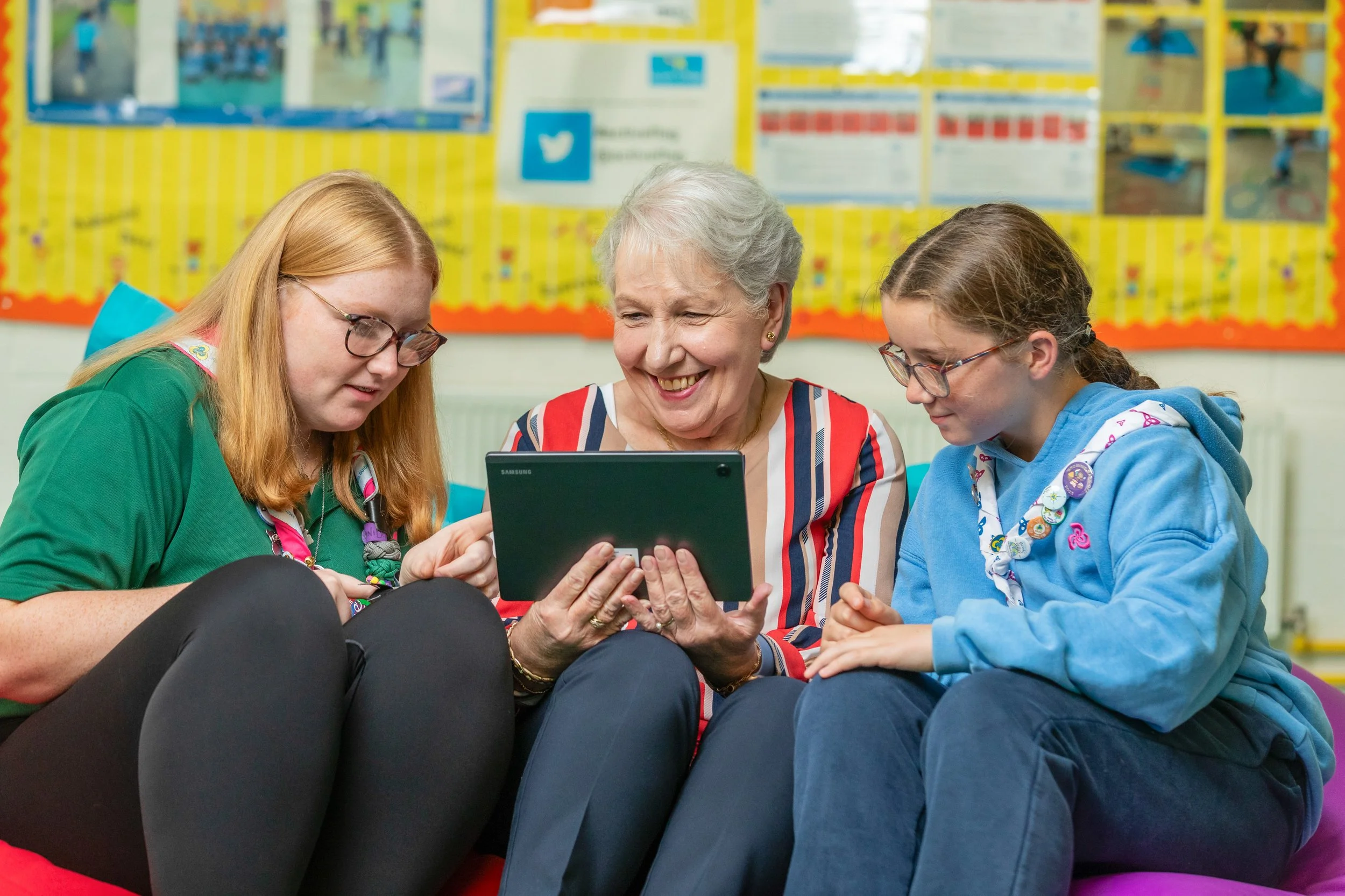 An older white woman sits in a classroom environment with two girls either side. They are smiling and looking at a device screen.
