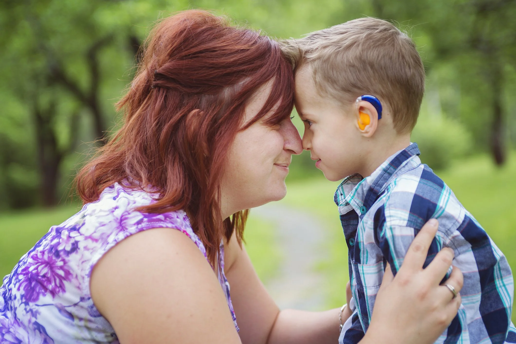 A white woman and a young boy wearing a hearing aid are nose to nose, smiling at each other in a park.