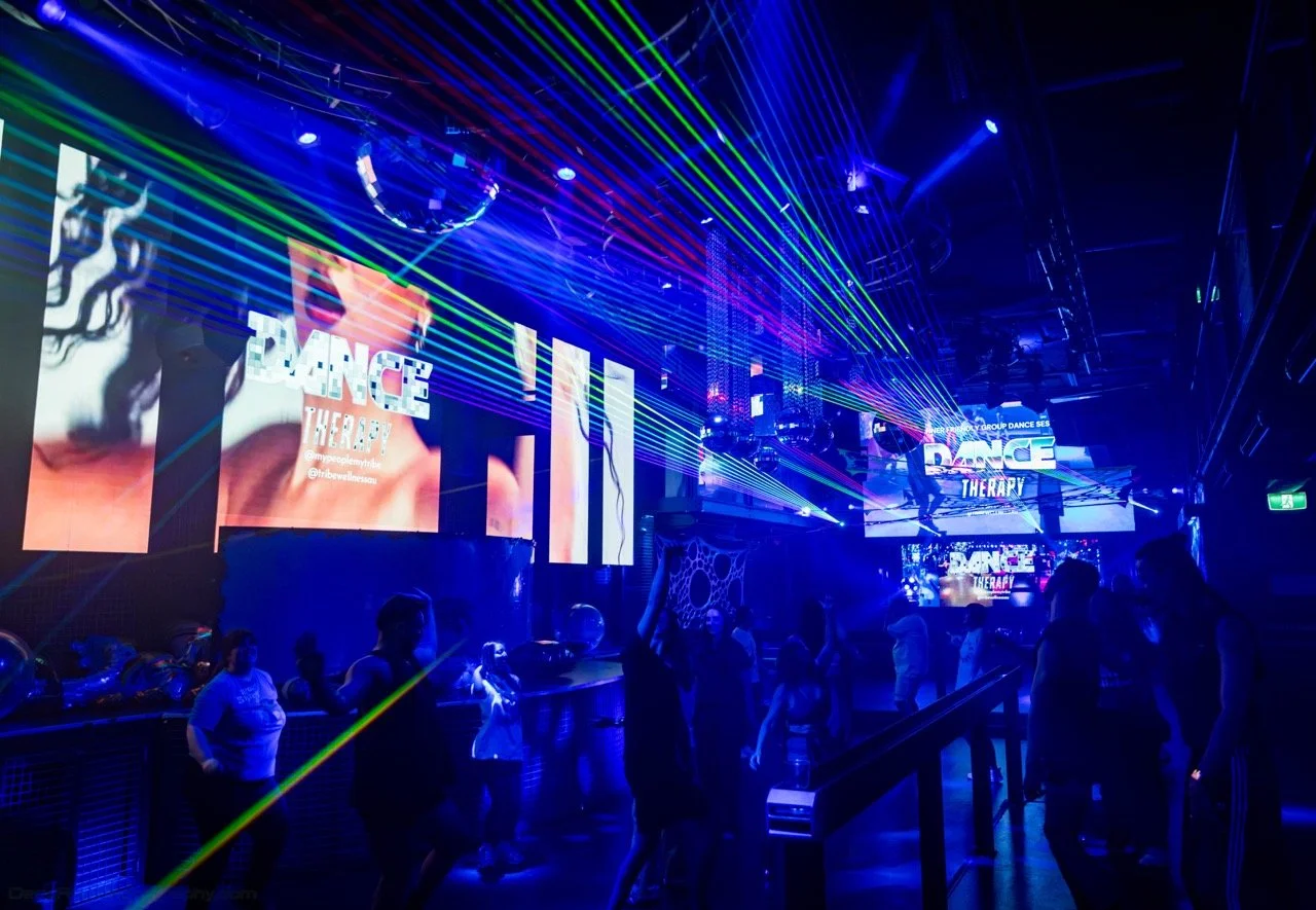 Dance Therapy nightclub scene with blue and green laser lights, large screens displaying Dance Therapy graphics and people dancing and connecting with community thanks to Tribe Wellness.