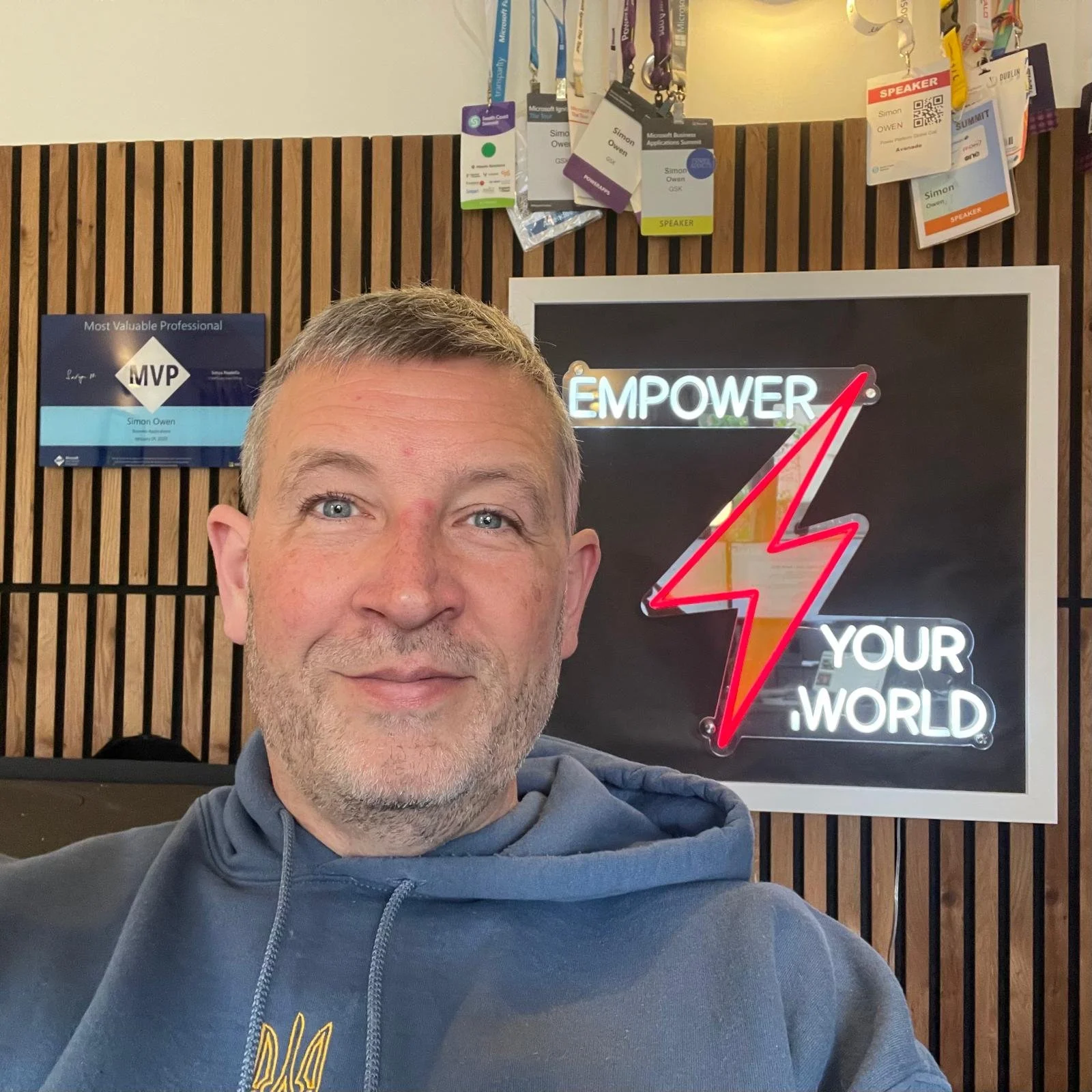 A man with short gray hair and blue eyes taking a selfie in front of a wooden wall with framed signs, one of which says 'Most Valuable Professional MVP' and another reads 'EMPOWER YOUR WORLD' with a lightning bolt symbol.