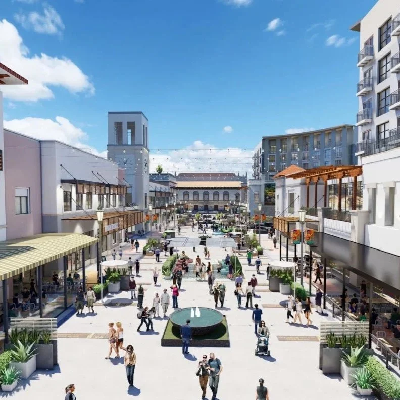 A lively outdoor shopping center with multiple levels, shops, and cafes. The wide pedestrian area is filled with people walking, talking, and sitting, with planters and decorative elements. Modern buildings surround the plaza, and there is a clock tower in the background under a partly cloudy sky.