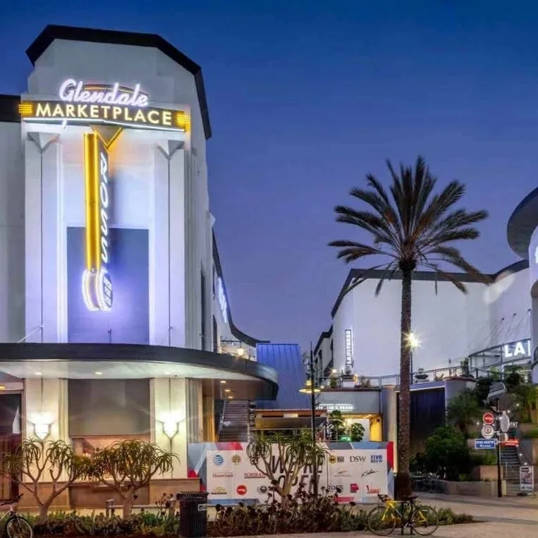 Nighttime view of Glendale Marketplace with a neon sign reading 'Glendale' and a vertical neon sign that says 'Theatre'. There is a tall palm tree in front, small trees, and a bicycle parked on the sidewalk.