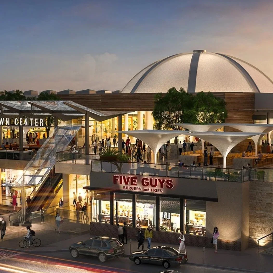 A shopping mall exterior at dusk with a dome-shaped roof, an outdoor seating area, a Five Guys burger restaurant, pedestrians, cars, and trees.