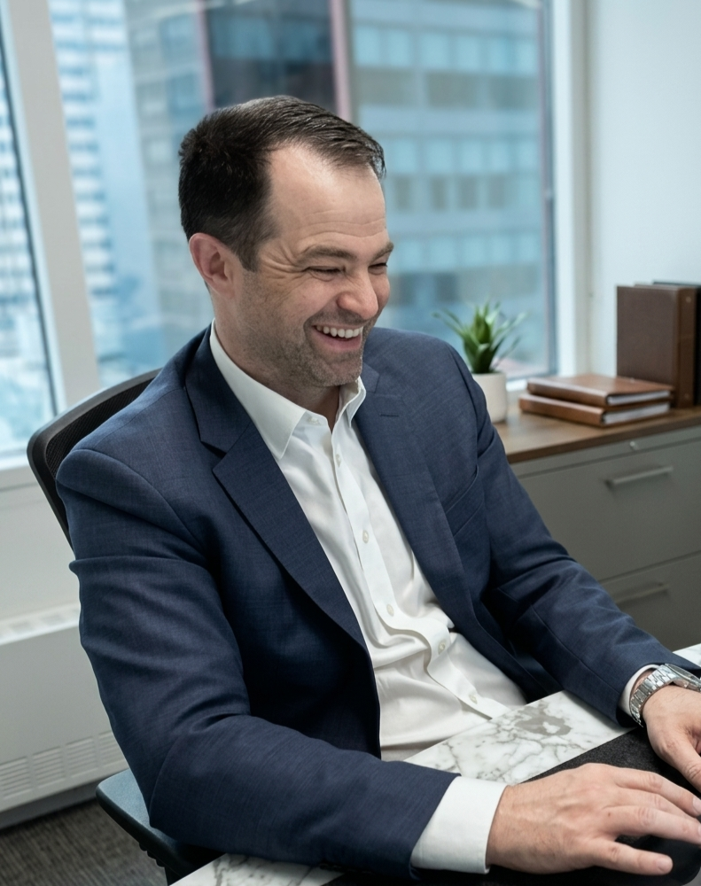 A man in a blue suit and white shirt sitting at a desk, smiling in an office with windows showing city buildings in the background.