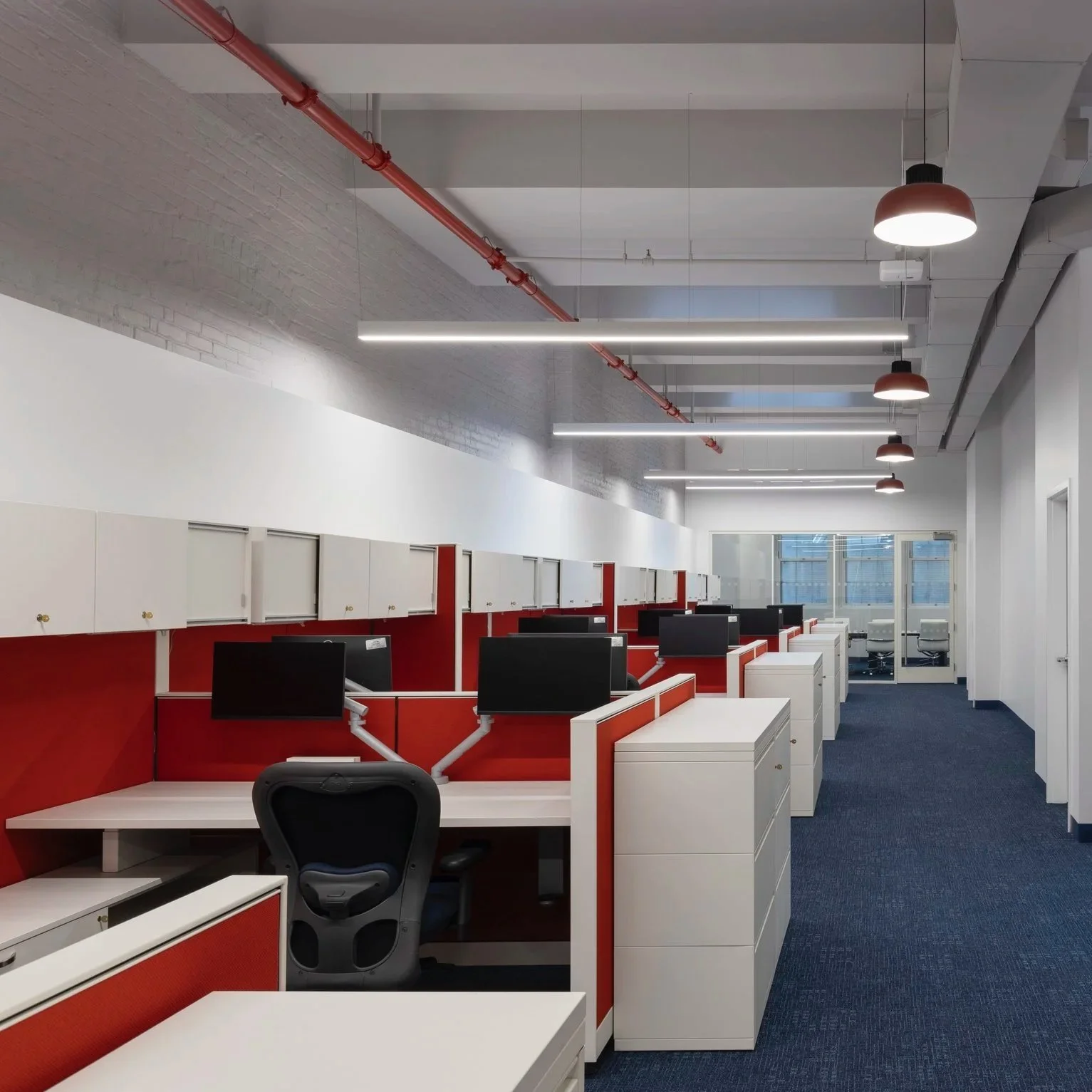 An office space with cubicles featuring red and white panels, black computer monitors, office chairs, and overhead lighting, with a view of windows and a glass door at the far end.
