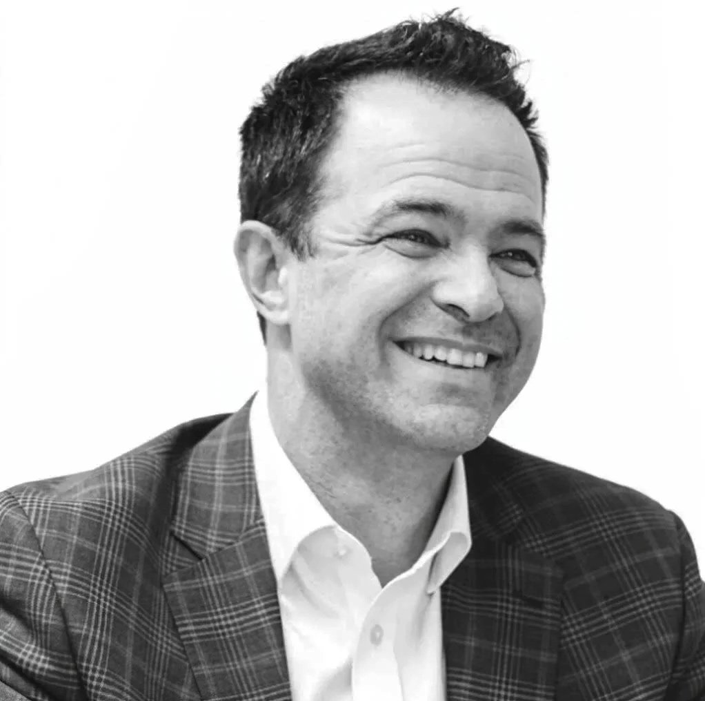 Black and white portrait of a smiling man with short dark hair, wearing a checkered blazer and white shirt.