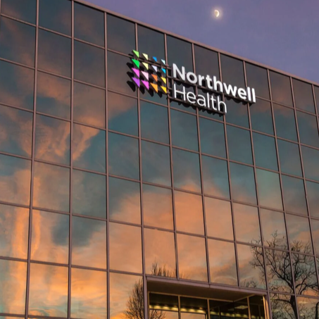 Exterior of Northwell Health building with reflective glass windows showing a sunset sky and partial moon.