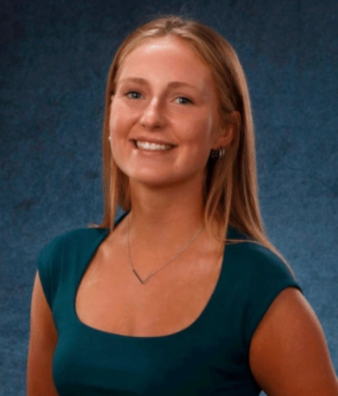 A woman with shoulder-length reddish-brown hair smiling, wearing a teal dress and a necklace, against a blue background.
