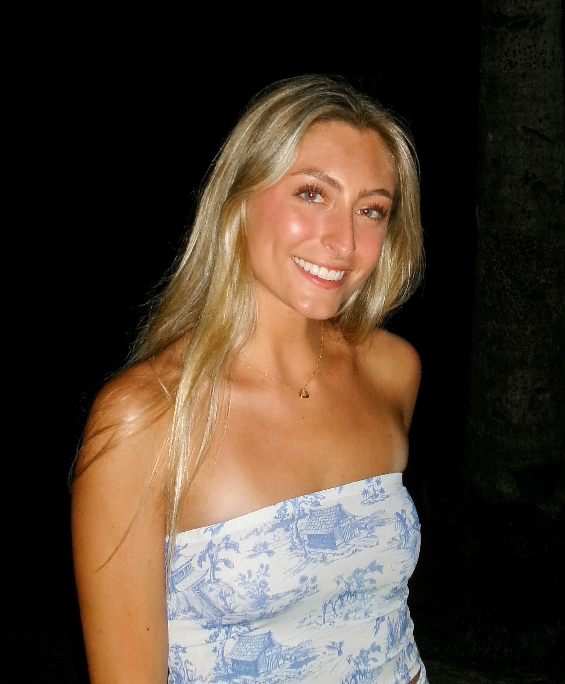 A young woman with long blonde hair smiling, sitting outside among green bushes, wearing a brown one-shoulder top.