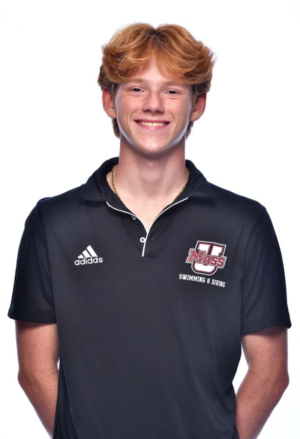 Smiling young man with red hair wearing a black athletic polo with University of Massachusetts logo and Adidas logo, posing against a plain white background.