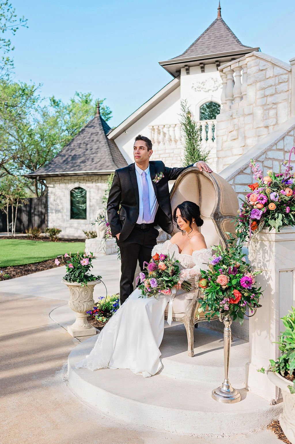 a bride and groom pose out front of castle-like wedding venue in an upholstered chair surrounded by flowers