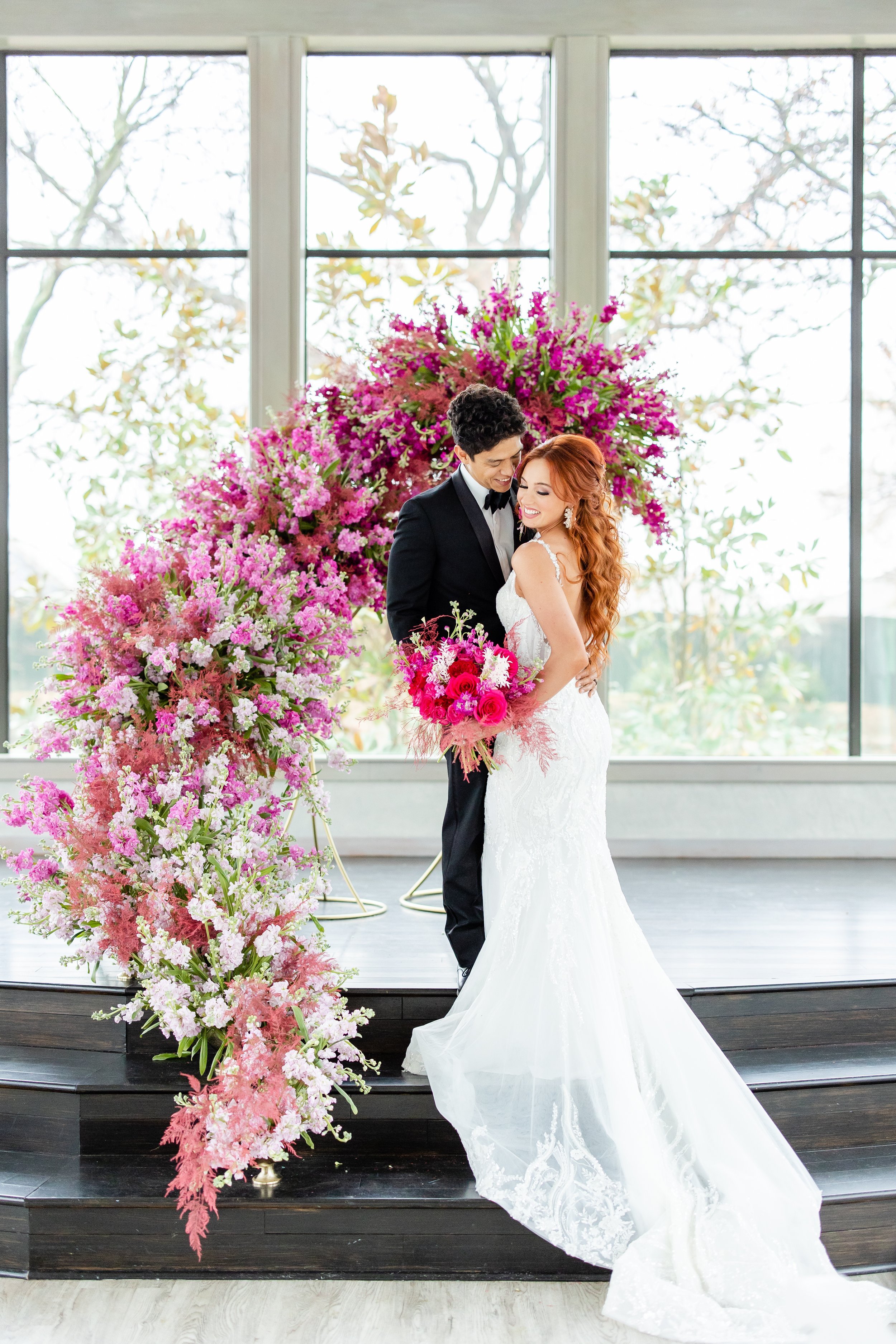 A bride and groom pose on an alter with a floral display behind them and huge windows in the background