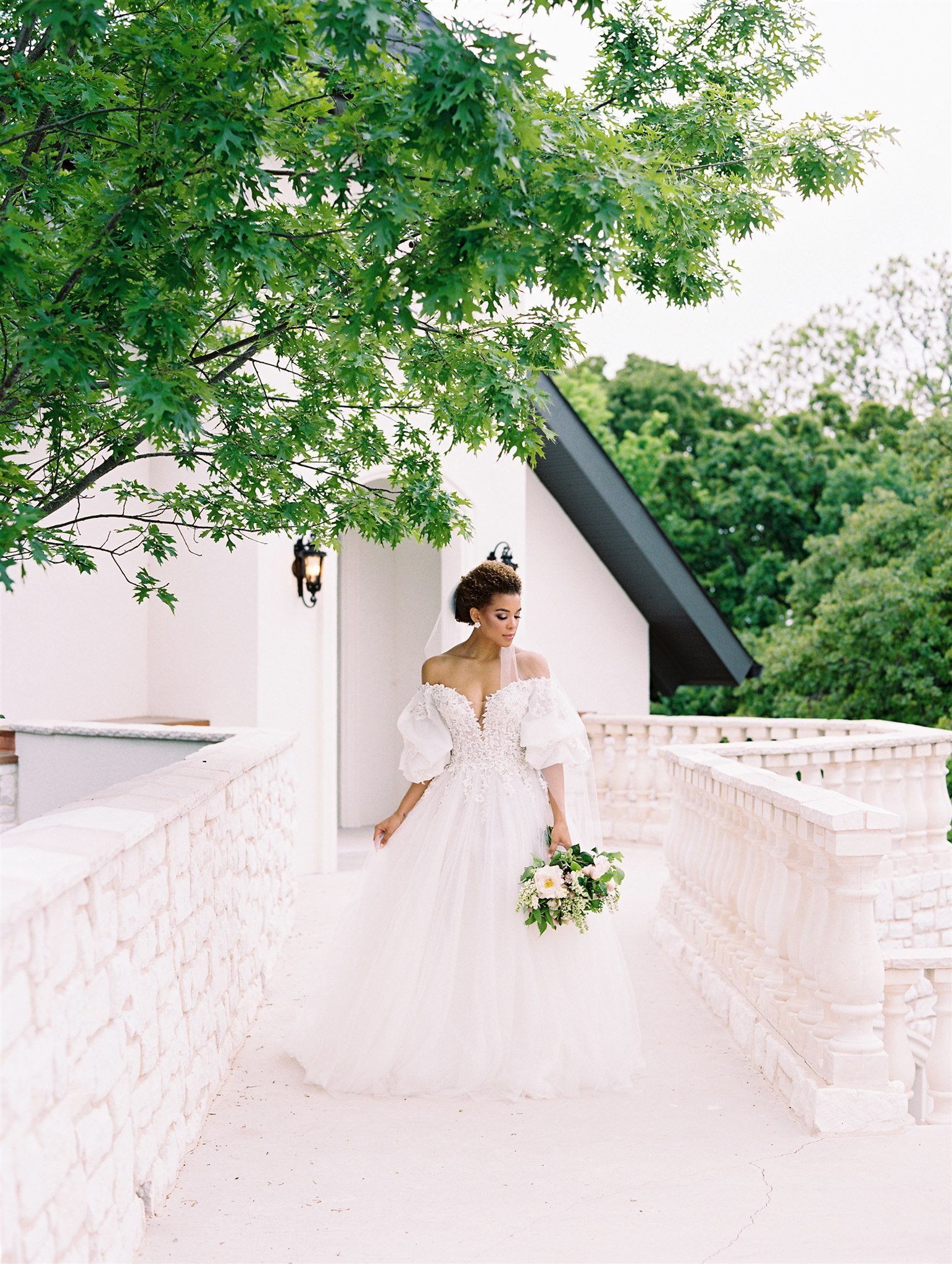 a bride walks through a stunning balcony in an off-the shoulder gown, her bouquet down at her side.