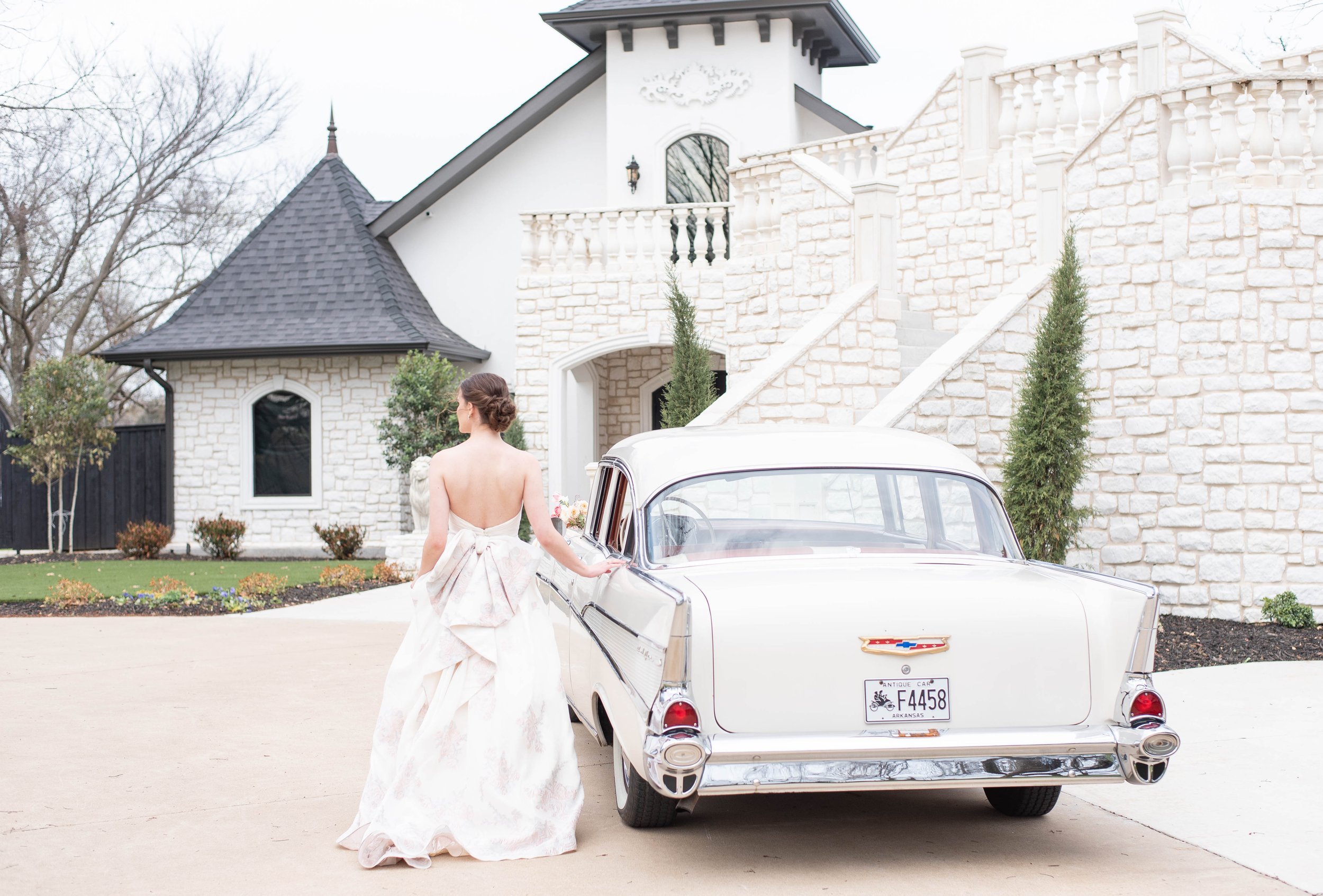 a bride walks toward a vintage car, her back to the camera otuside of a grand staircase.