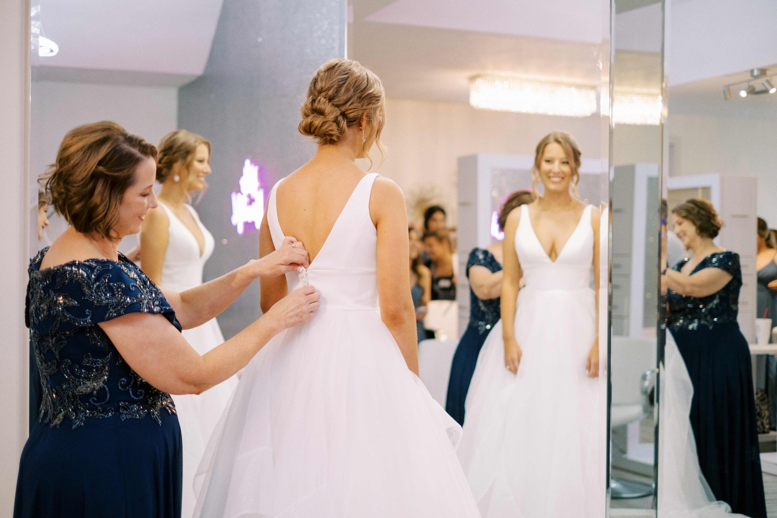 A mother helps her daughter into her wedding gown, bridesmaids look on and smile