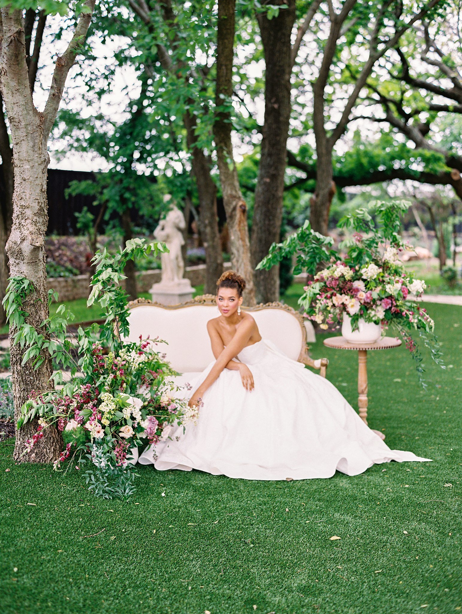 A bride sits outside on an upholstered couch, surrounded by flowers, her arms crossed over her lap