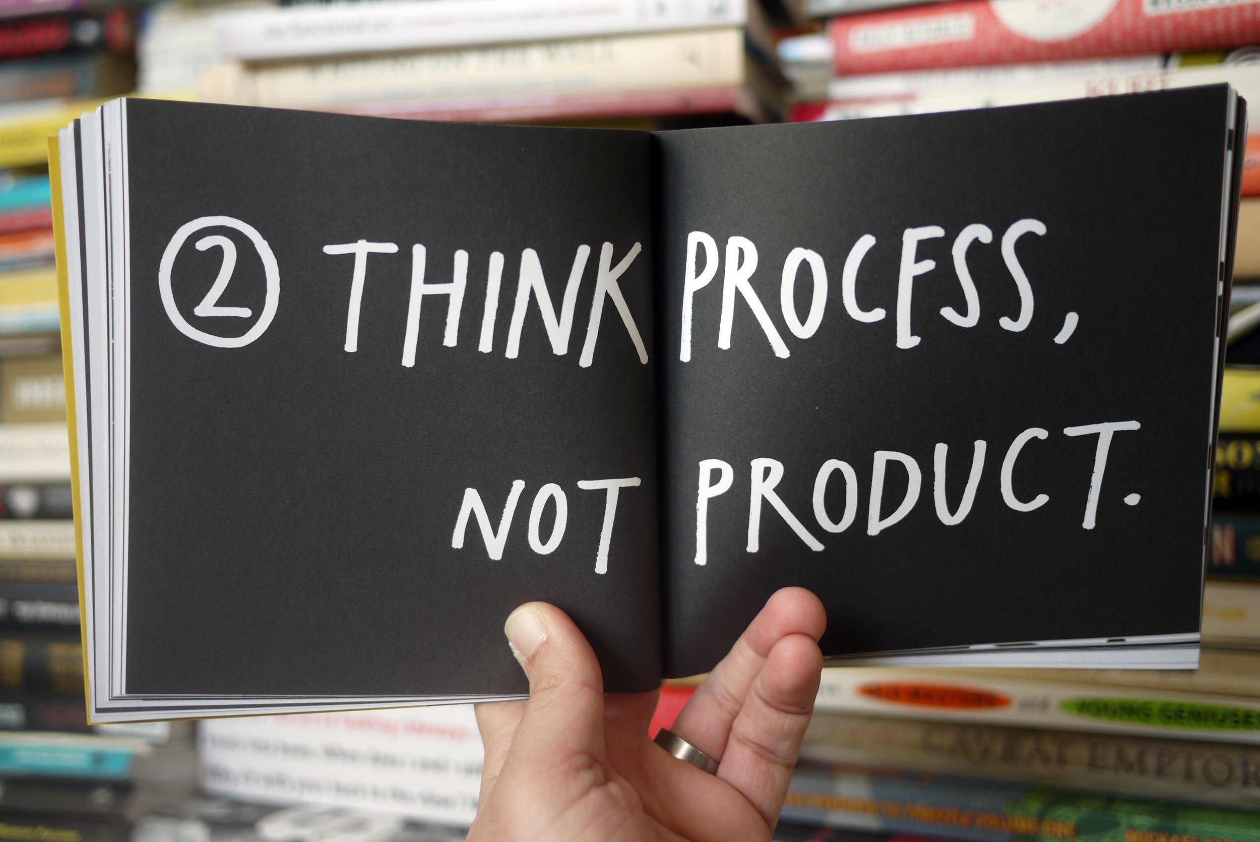 A hand holding open the book "Show Your Work" to a page with the words "Think process, not product"