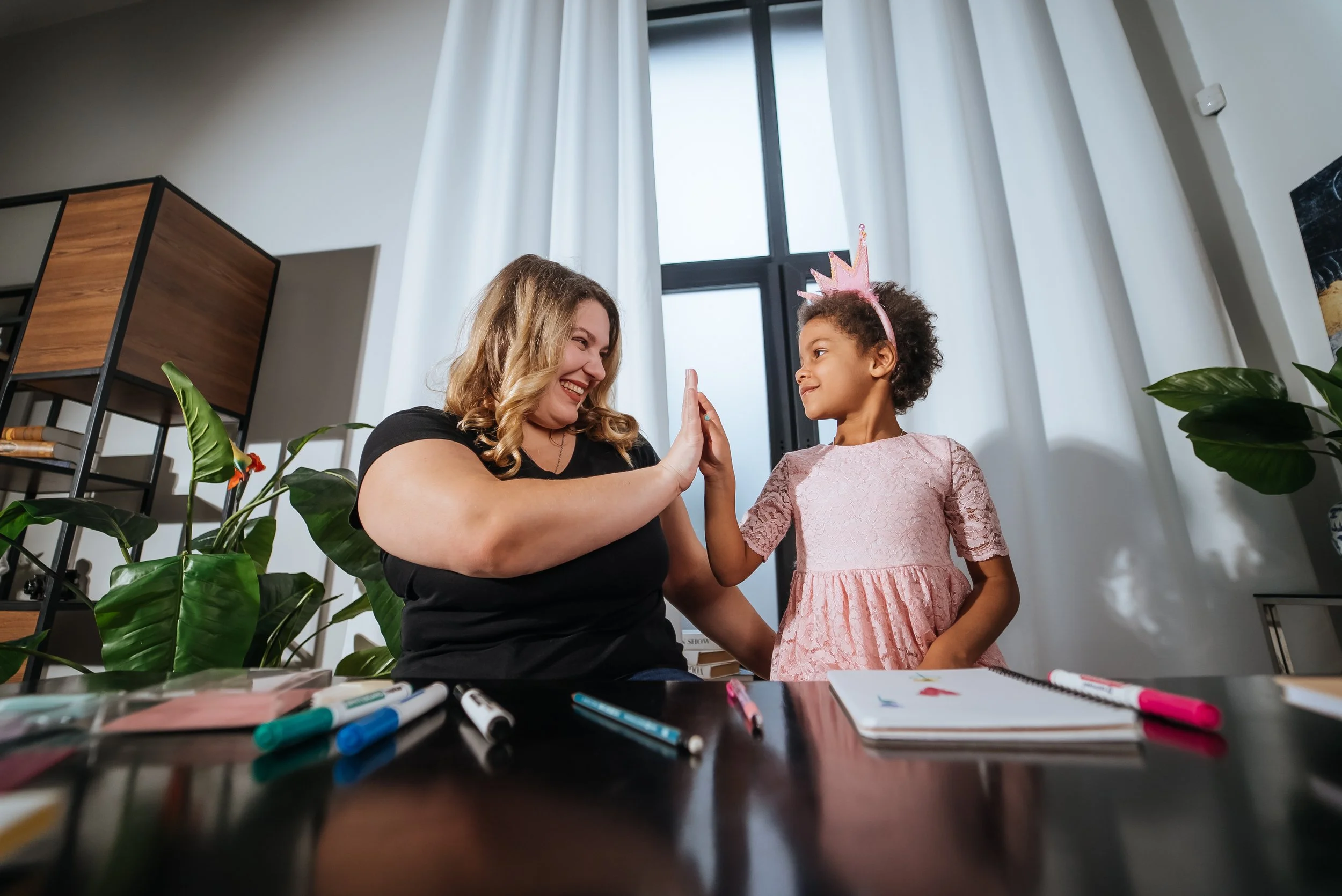 A woman and young girl giving each other a high five indoors, with drawing supplies and notebooks on the table in front of them, and a plant and window behind them.