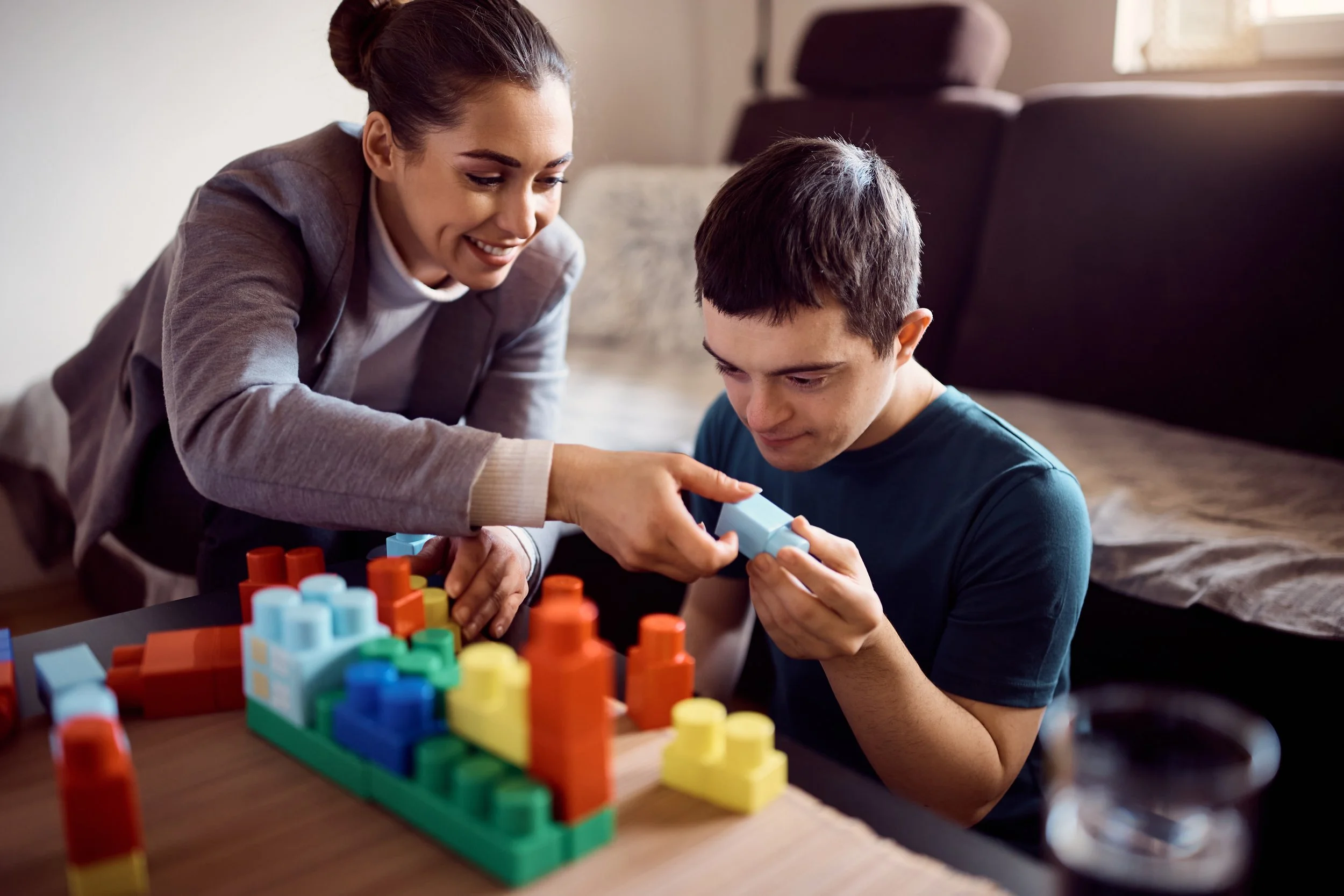 A woman and a young man playing with colorful building blocks at a table in a living room.
