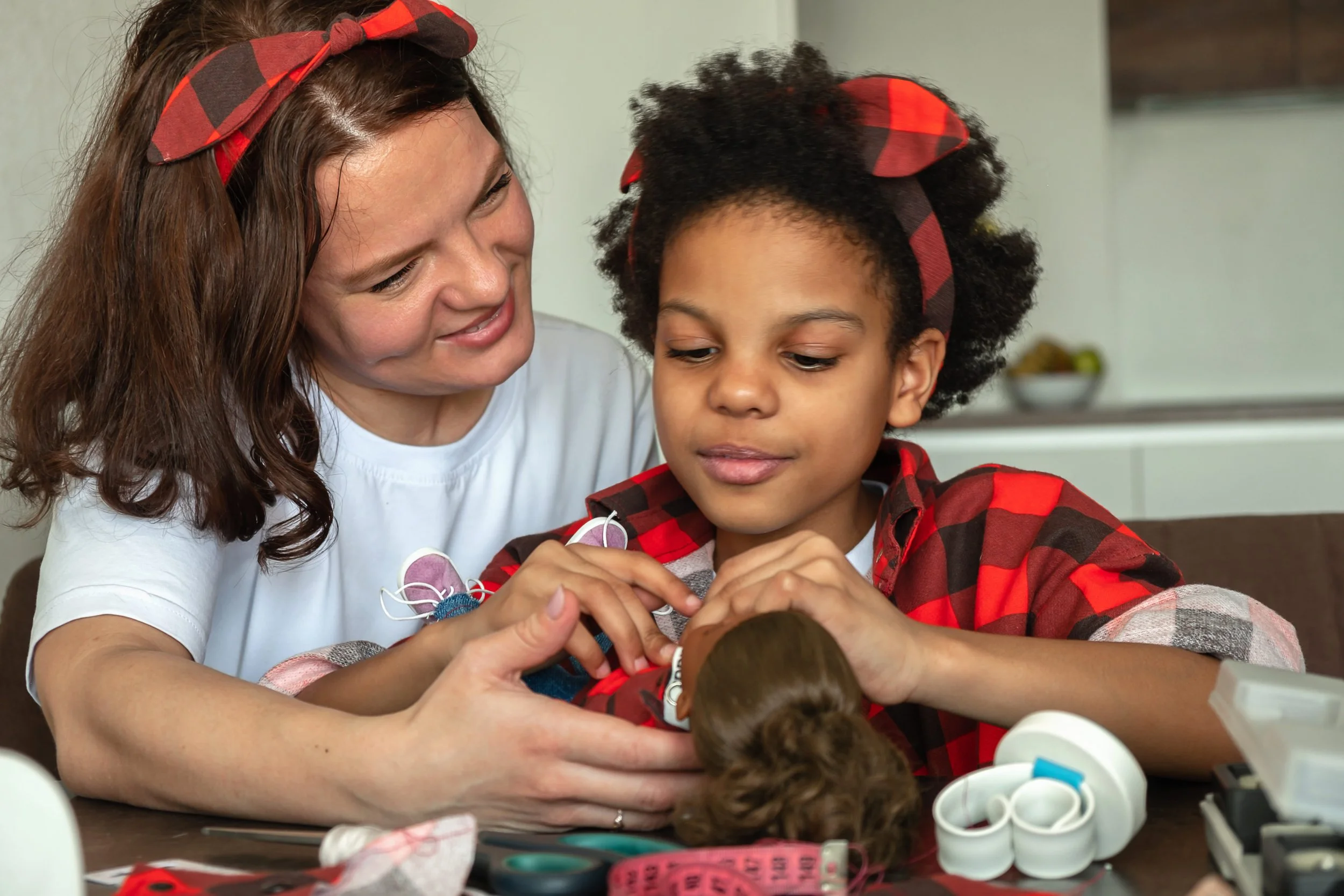 A woman and a boy, both wearing red and black checkered headbands and shirts, are working together on a craft project at a table. The woman is smiling and helping the boy, who is focused on the craft materials in front of him.