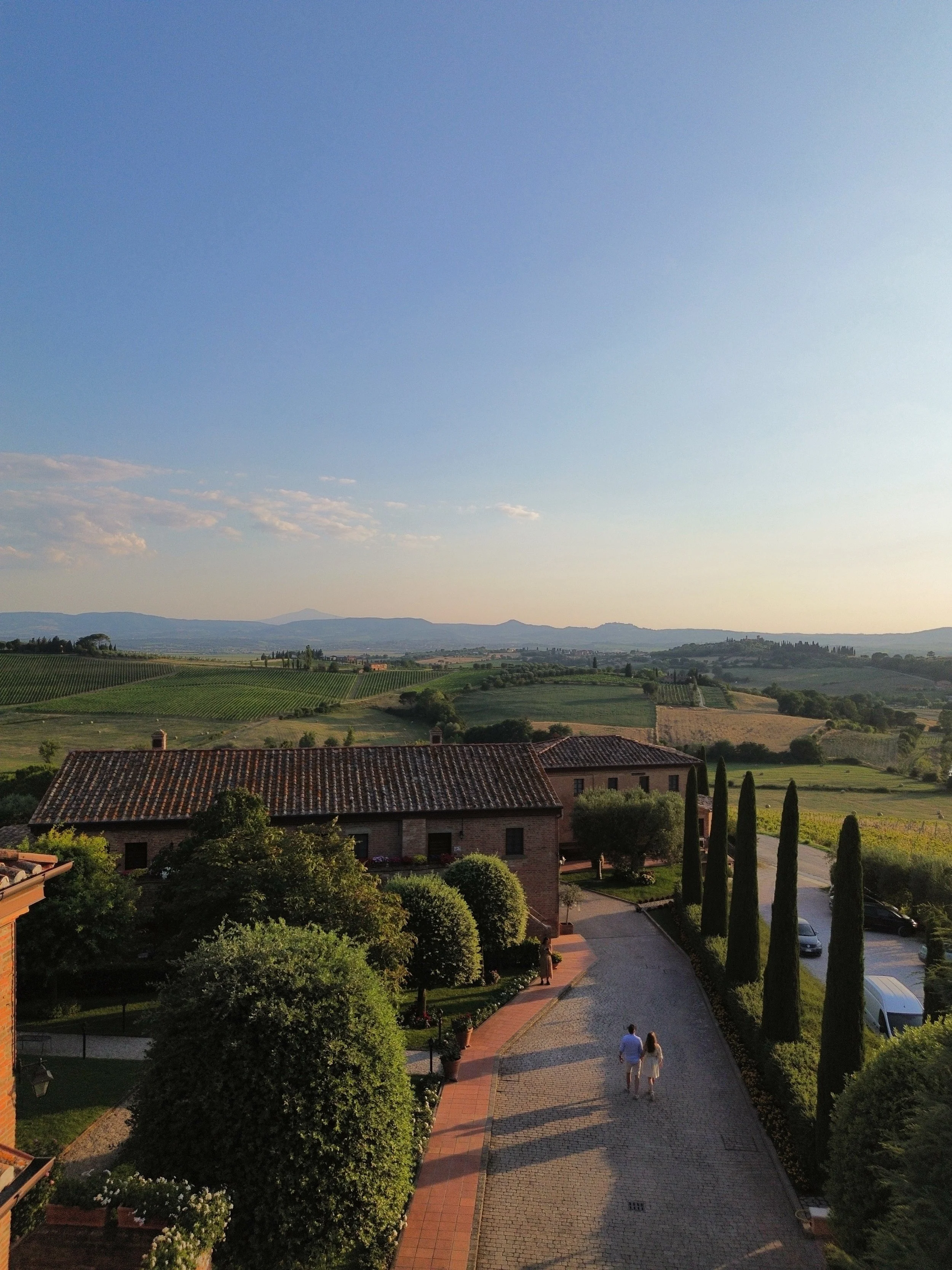 Scenic view of a countryside with rolling hills, vineyards, and distant mountains under a clear blue sky. A cobblestone pathway runs through a landscaped area with trees and parked cars, with two people walking along it.