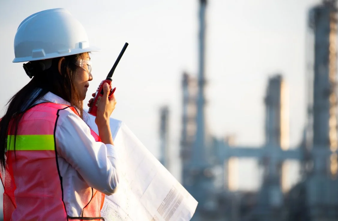 A woman wearing a white hard hat, safety glasses, a pink safety vest, and a white shirt holding a walkie-talkie and a large blueprint at an industrial construction site with cranes in the background.