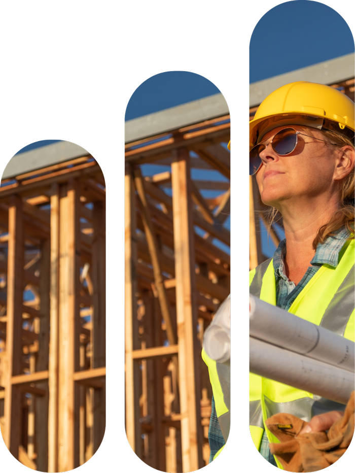 A female construction worker wearing a yellow hard hat and sunglasses holds blueprints at a wooden construction site during daytime.