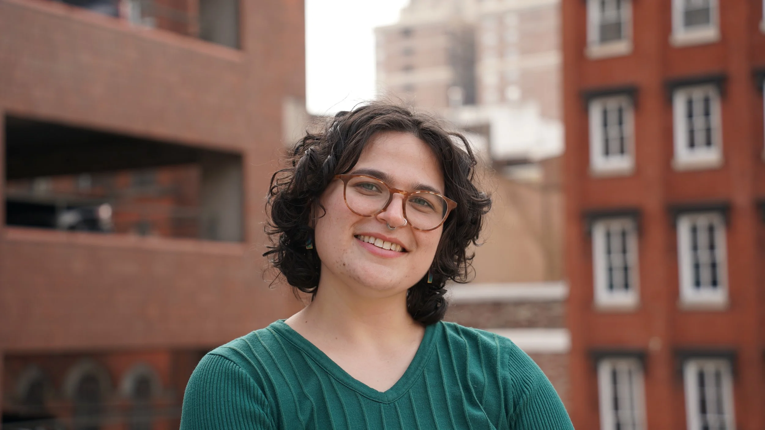 A person with curly dark hair and glasses outdoors, wearing a denim shirt, holding colorful ribbons, with trees in the background.