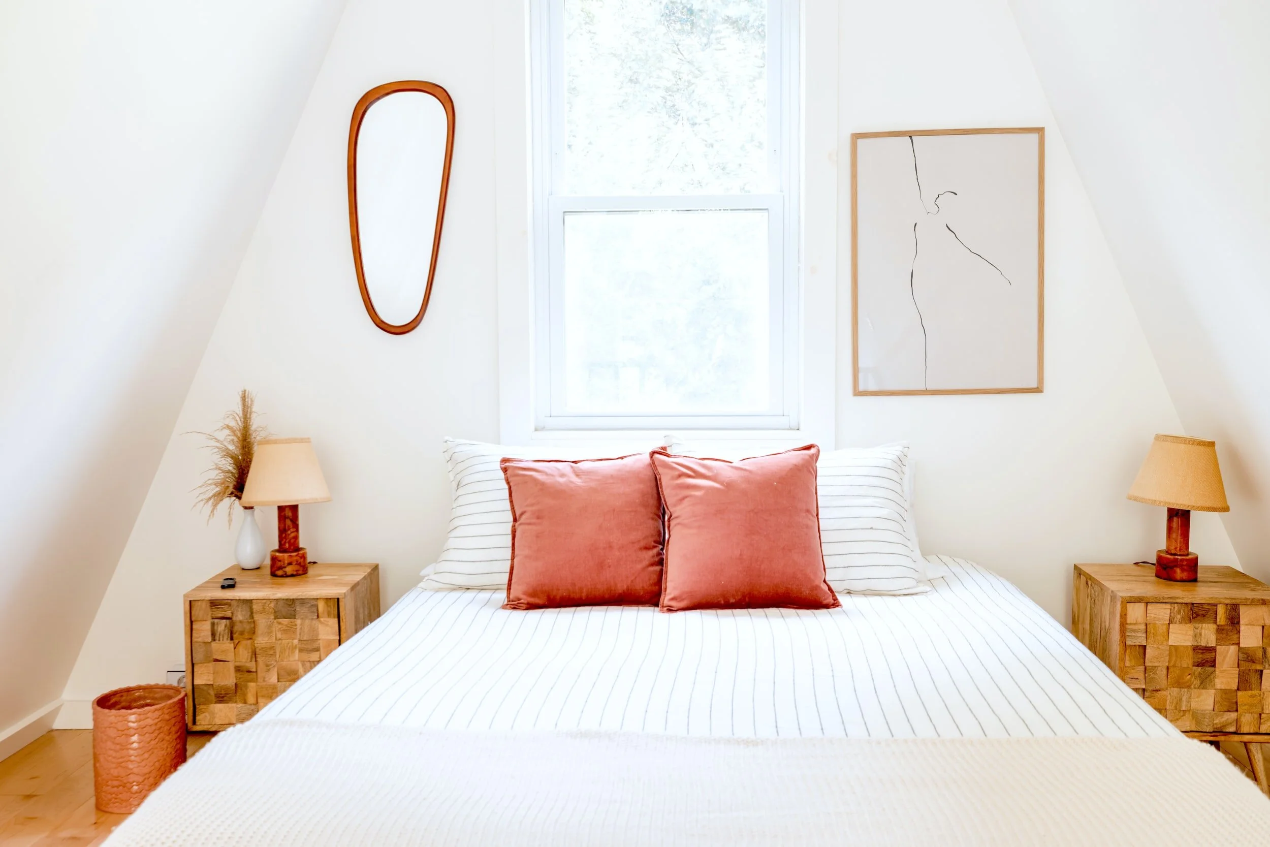 Minimalist bedroom with a white bed, two red pillows, wooden side tables, lamps, a mirror, and abstract wall art.
