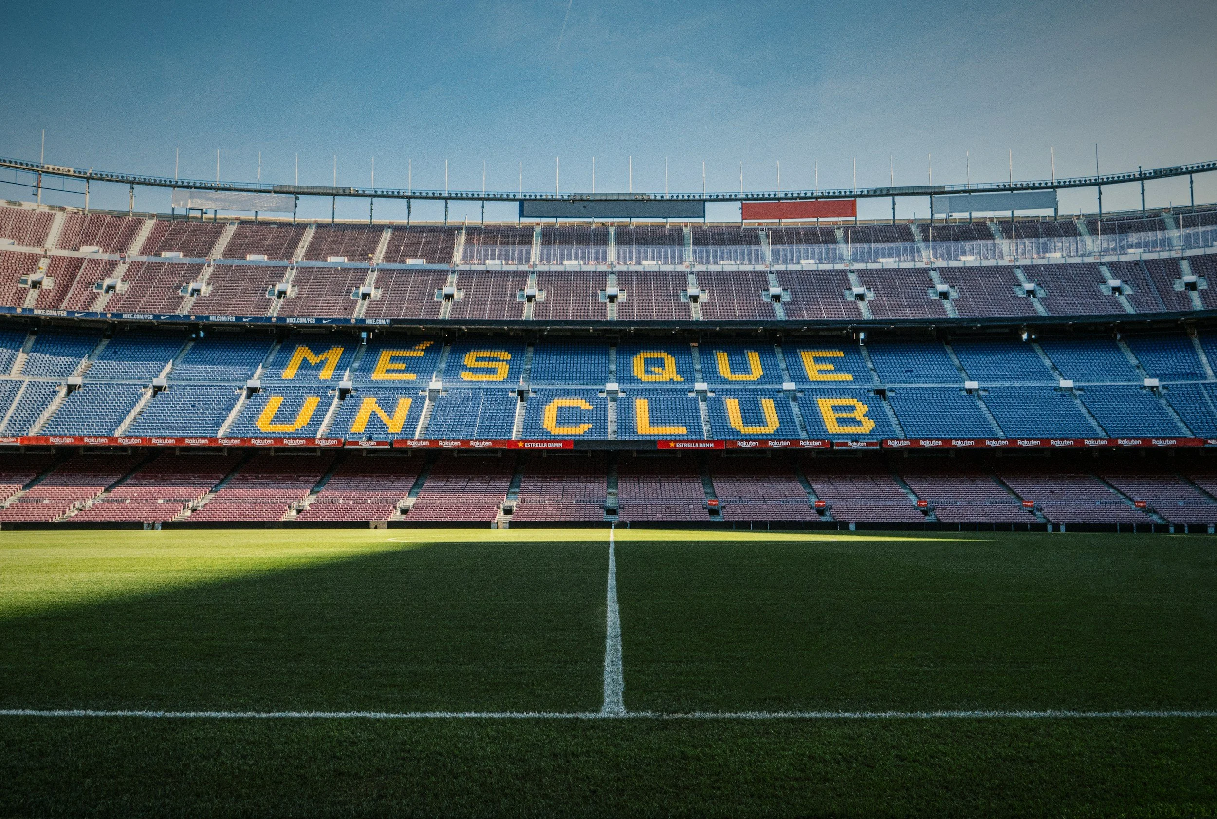 Empty soccer stadium with blue seats displaying yellow letters spelling 'MÈS QUE UN CLUB', and a well-maintained grassy field under a clear blue sky.