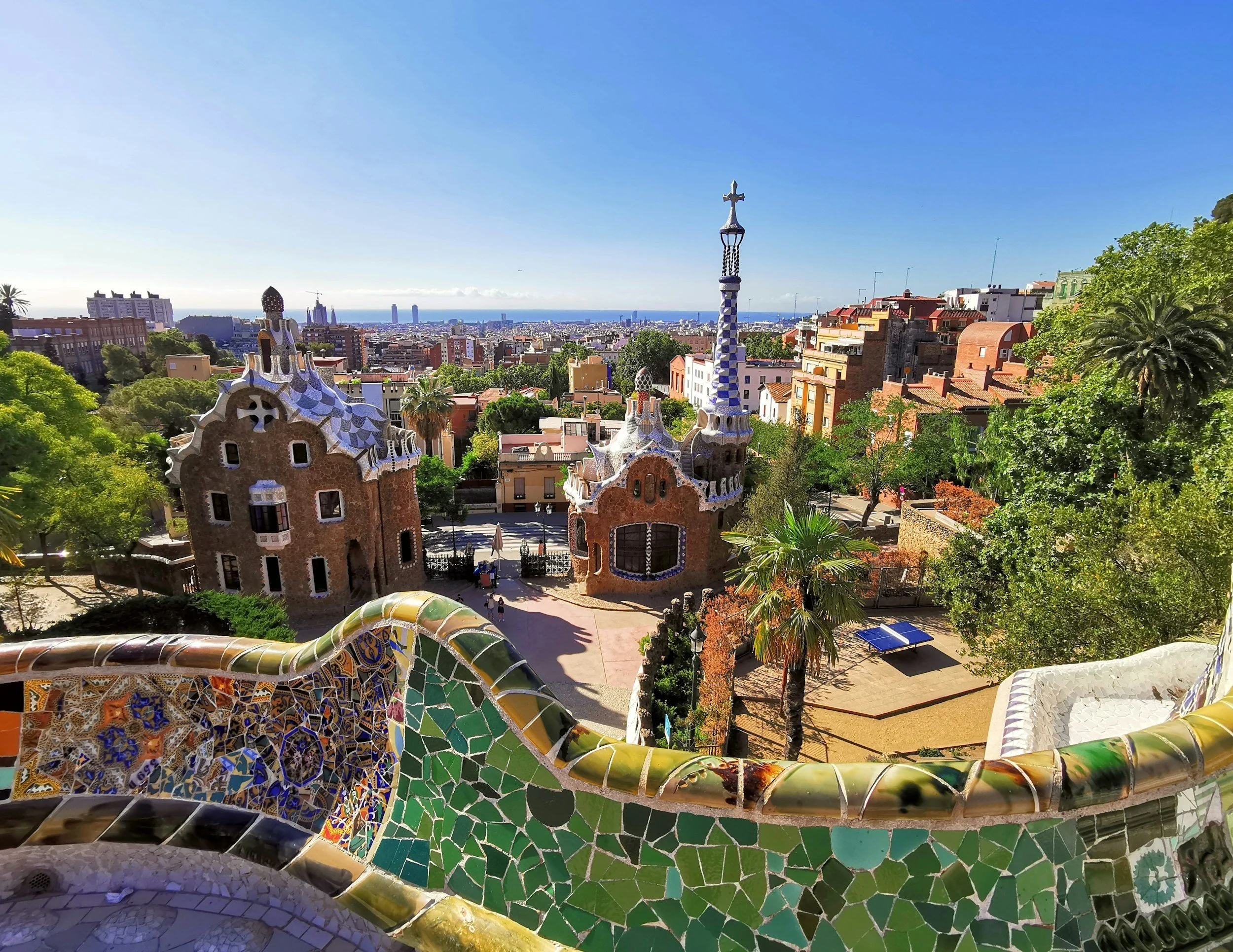 Colorful mosaic dragon sculpture with the cityscape of Barcelona in the background, including buildings, trees, and the sea under clear blue sky.