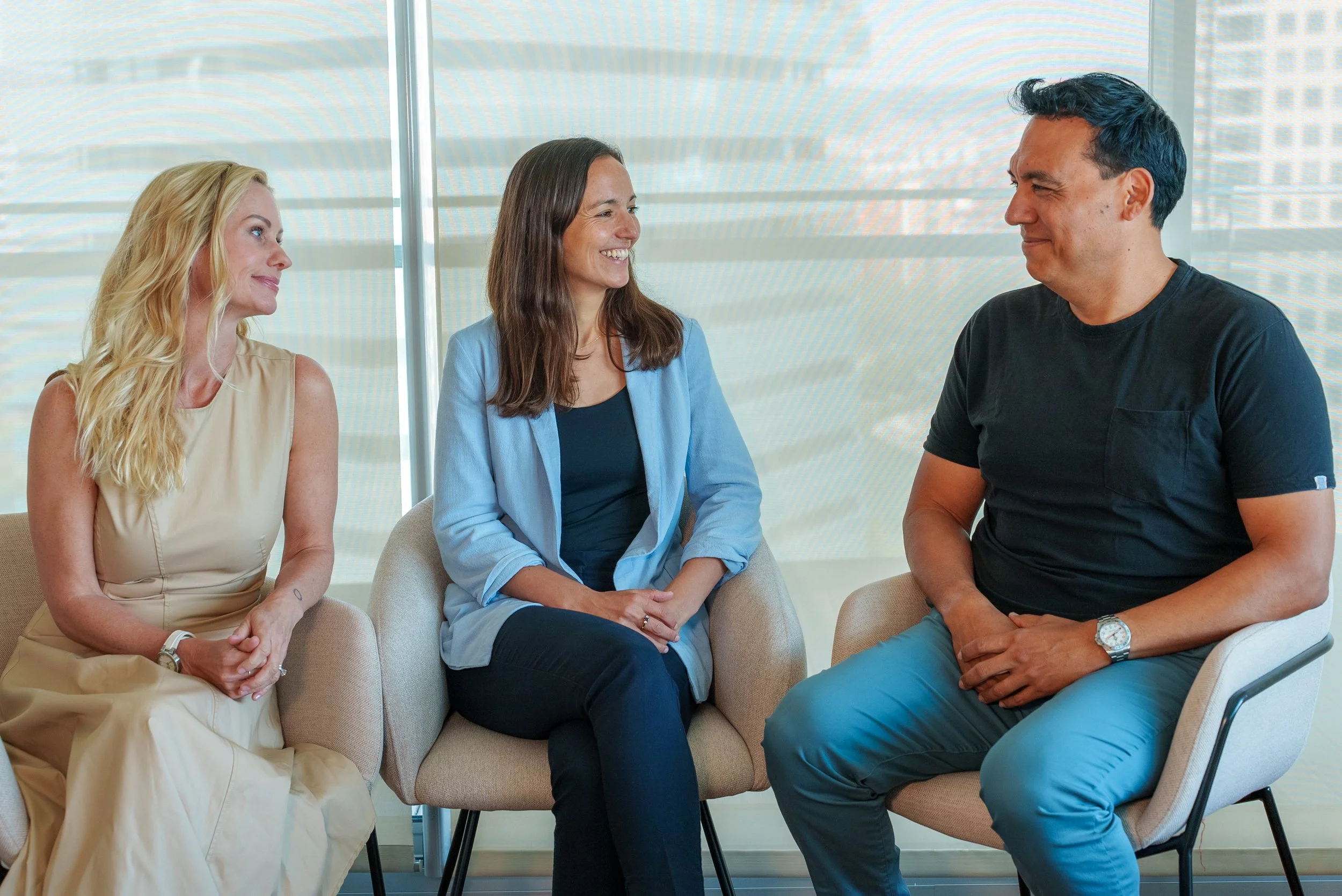 Lucy Mills, founder of Ready Sport Global, with Magdalyn Miller COO and Hector Dominguez CTO, on chairs, engaged in conversation in an office setting, with large windows and cityscape view in the background.