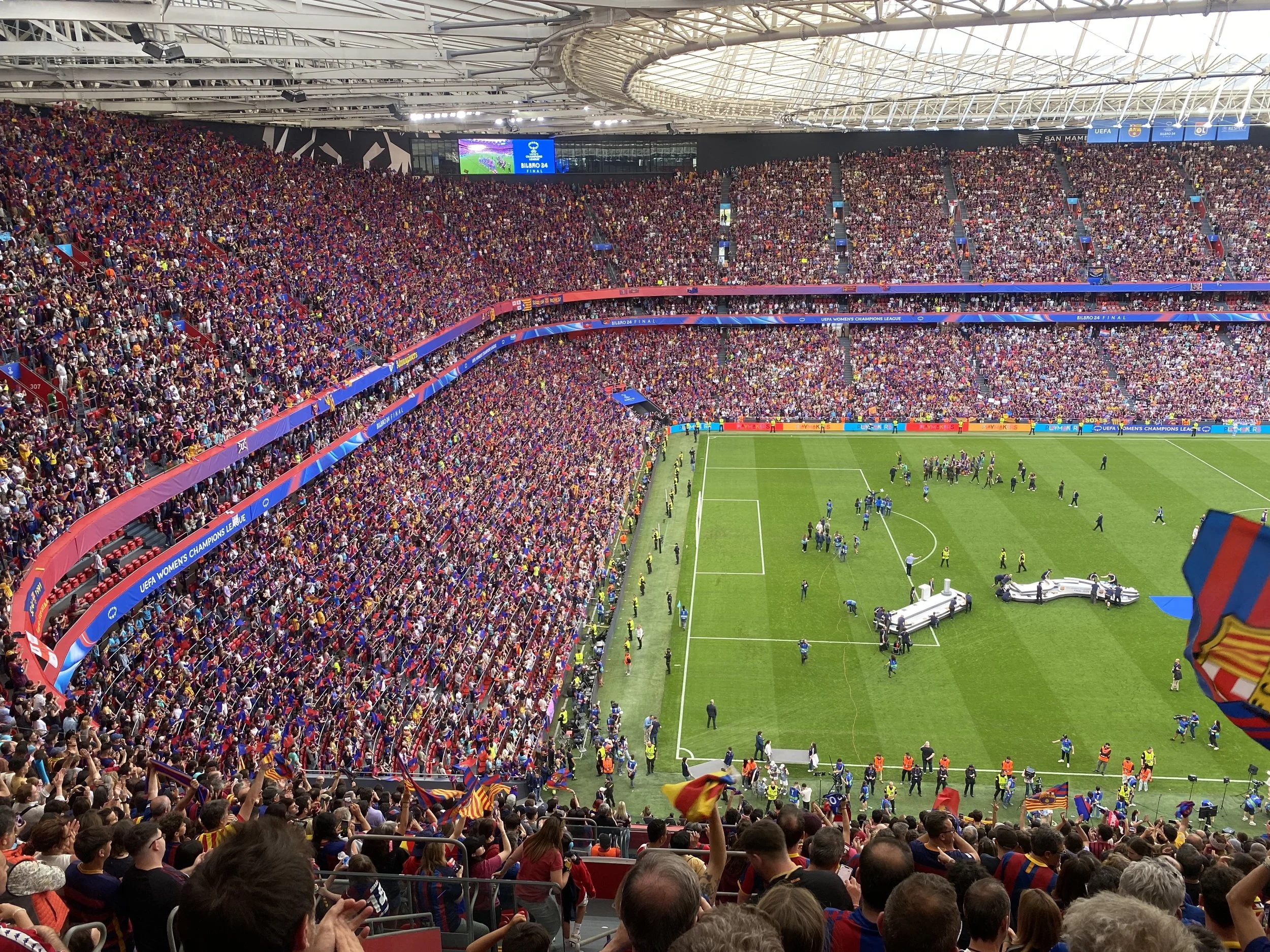 A crowded stadium filled with fans during a soccer match, many of whom are waving flags and wearing team colors. The field is visible in the lower right corner, with players and officials present. Large screens and banners are displayed around the stadium.