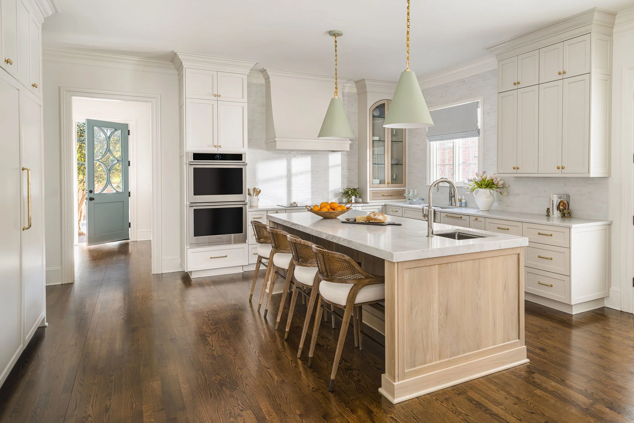 Transitional kitchen design in a Charlotte, NC home featuring a light oak island with woven cane barstools, custom white cabinetry, sage green pendant lights, and a classic arched glass pantry cabinet