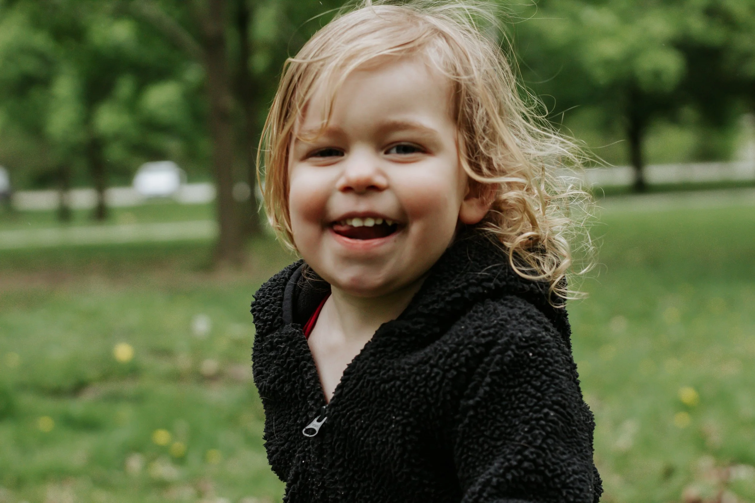 Close-up of a young child with curly blonde hair smiling outdoors in a park with blurred trees and grass in the background. Family photos at elgin park uxbridge ontario