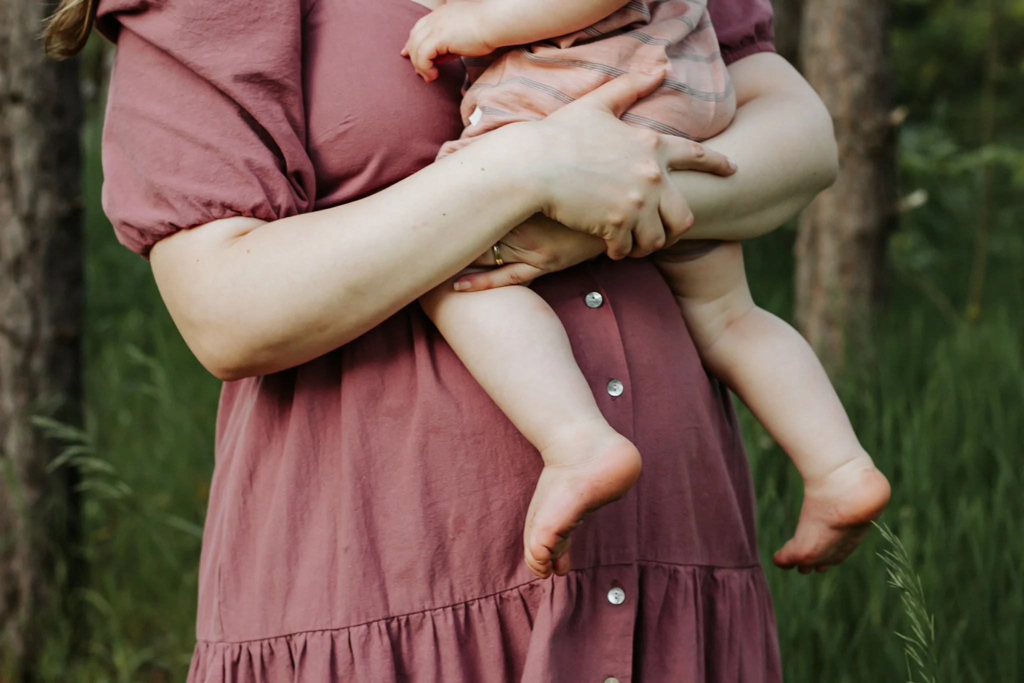 A woman in a pink dress holding a baby in her arms outdoors in a wooded area. maternity photos Uxbridge ontario