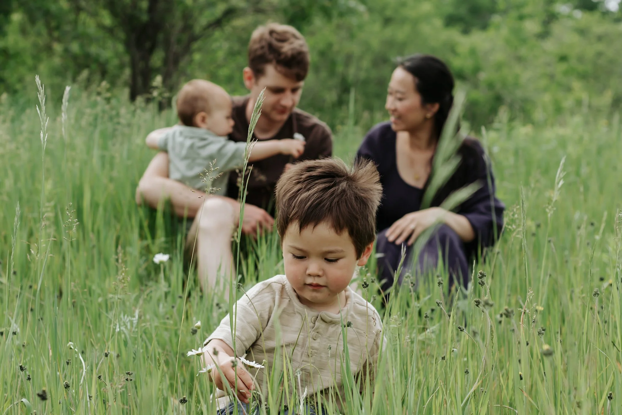 A family consisting of two children and two adults sitting and playing in a grassy field surrounded by trees.