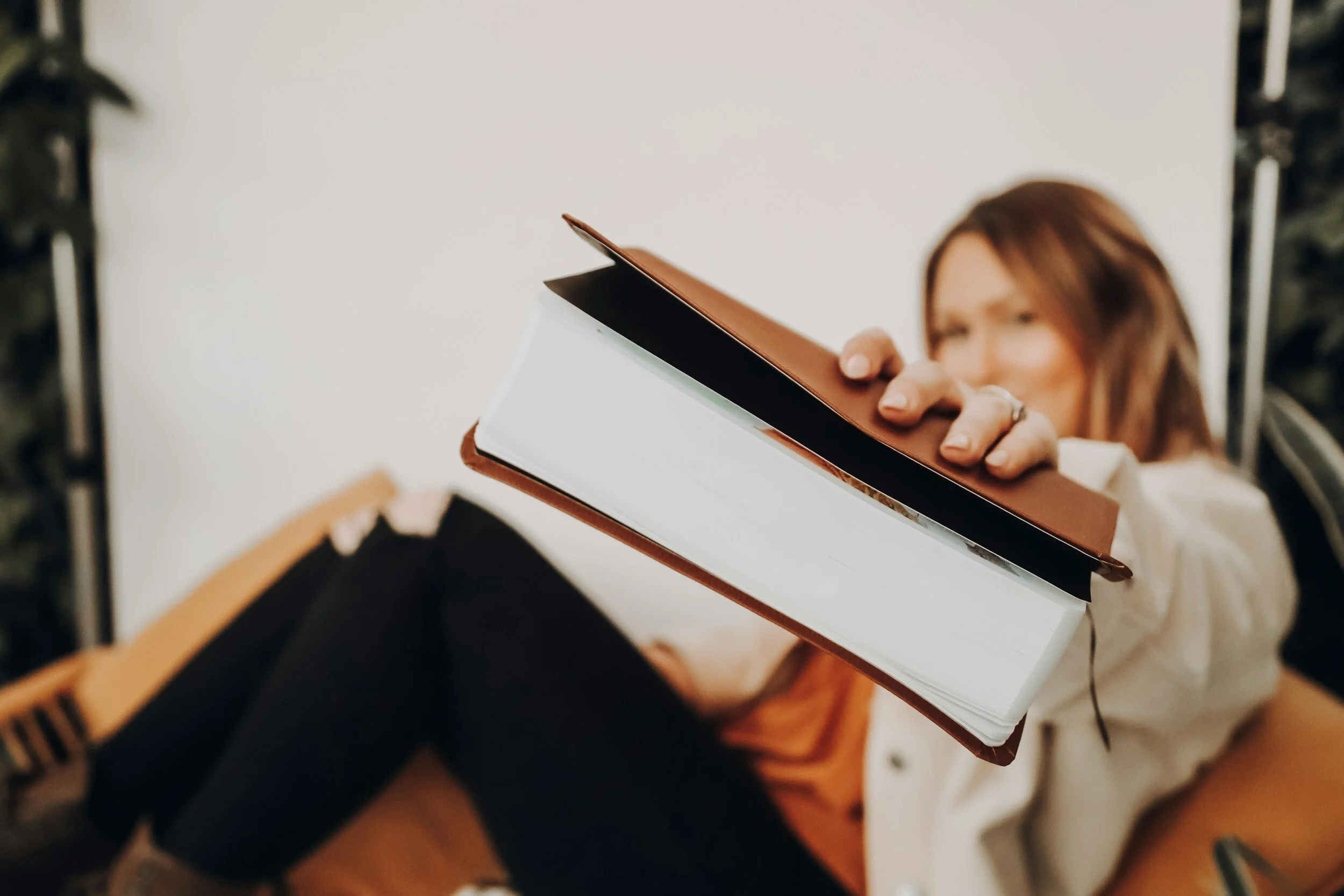 A woman with red hair holds a brown and white leather-bound notebook toward the camera. She is seated on an orange chair, wearing a white shirt and black ripped jeans. The background is plain and blurred.