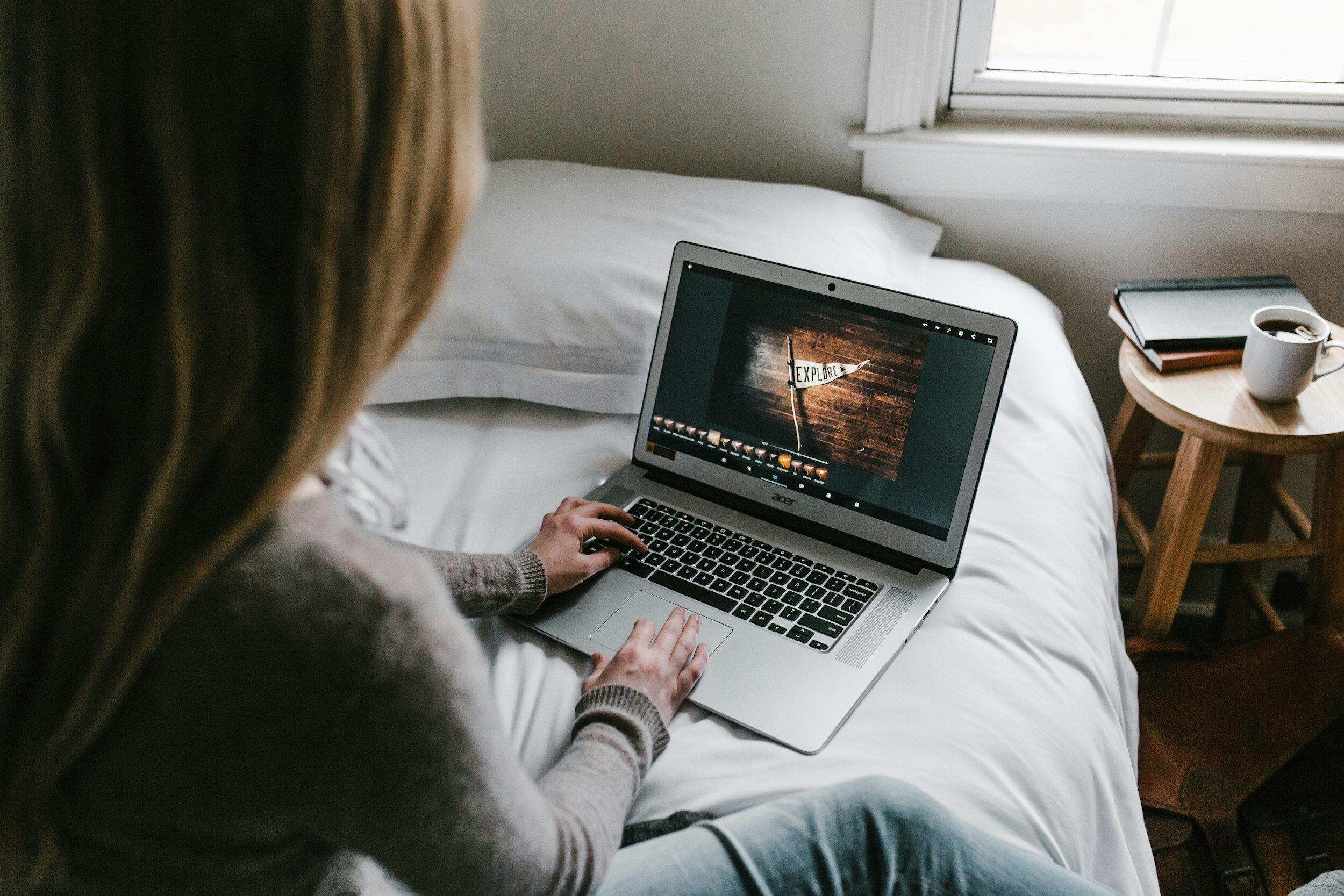 Person sitting on a bed using a laptop with a photo editing program open. The laptop screen shows a sign that says 'EXPLORE' on a wooden background. Beside the bed, there's a small round table with a coffee mug, a closed notebook, and a book or journal. The room has a window with natural light.